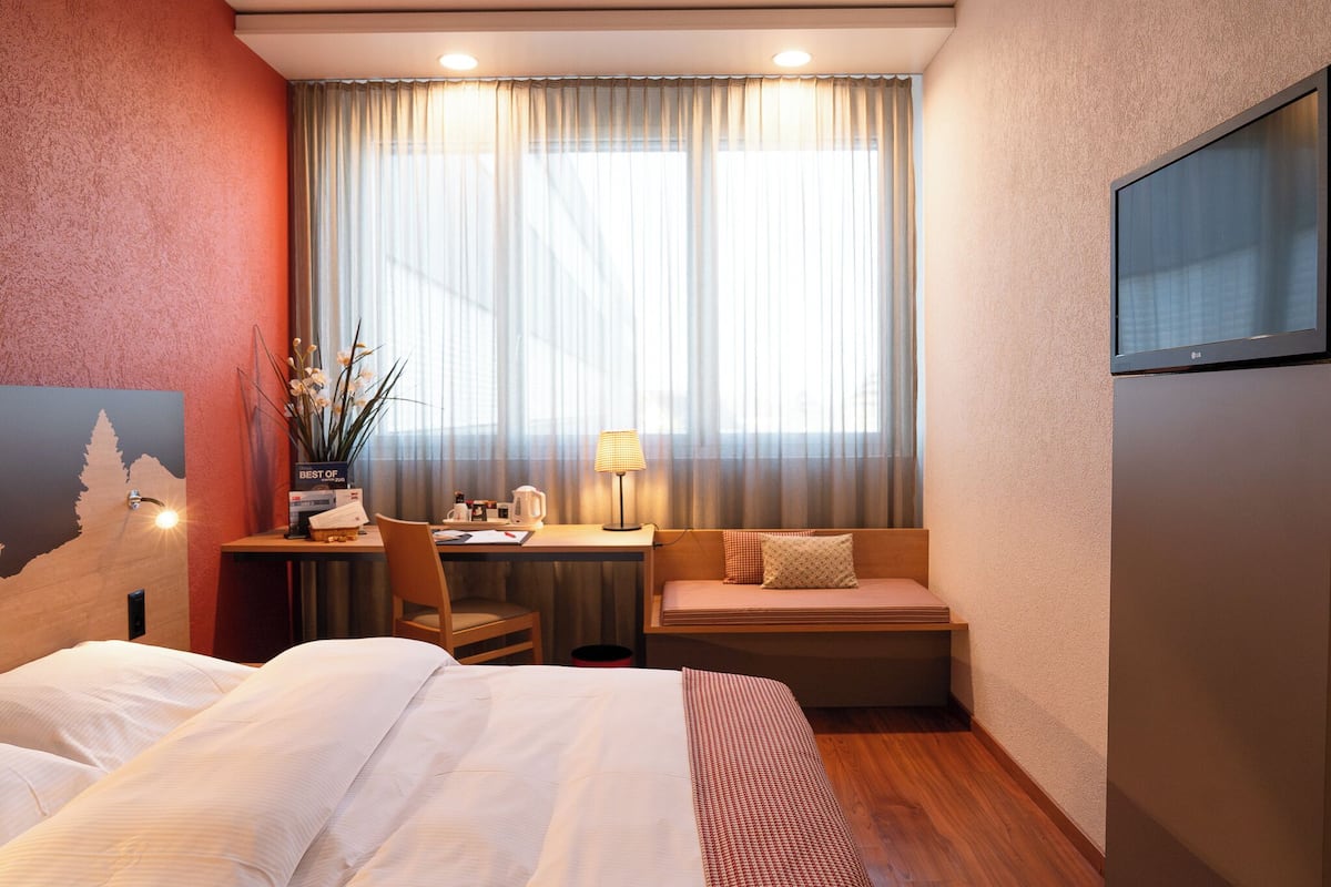 A hotel room with a bed, a desk with a lamp, a chair, a small table, and a window with curtains.