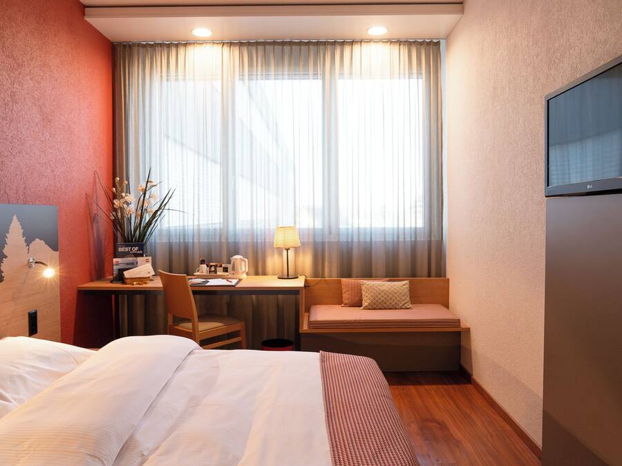 A hotel room with a bed, a desk with a lamp, a chair, a small table, and a window with curtains.