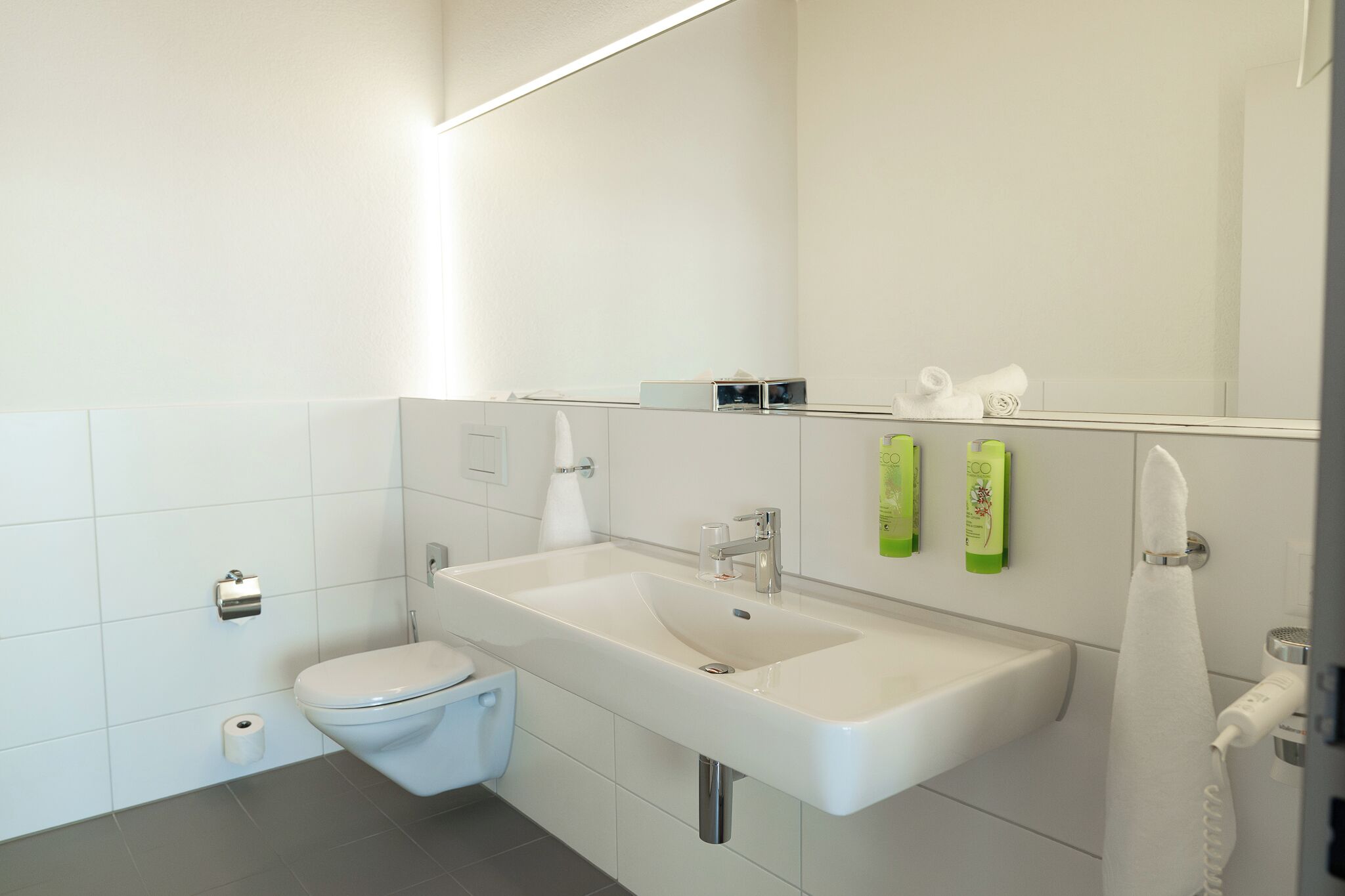 Une salle de bain moderne avec des toilettes blanches, un grand lavabo et un miroir au-dessus.