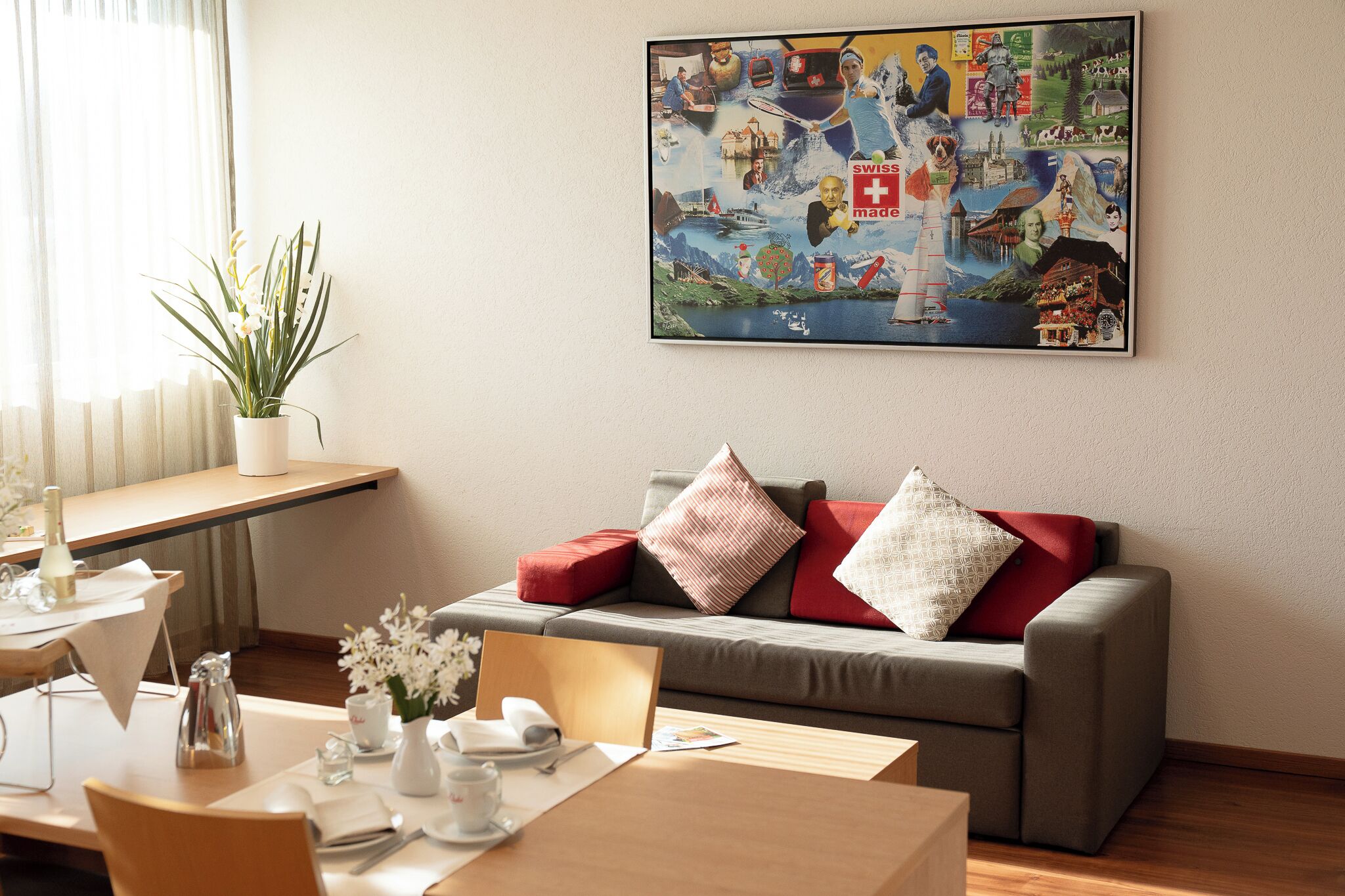 A modern hotel room with a dining area, a sofa with red and white cushions, and a large painting on the wall depicting Swiss landscapes and landmarks.