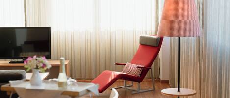A modern hotel room with a red lounge chair, a small round table, a TV, and a floor lamp.