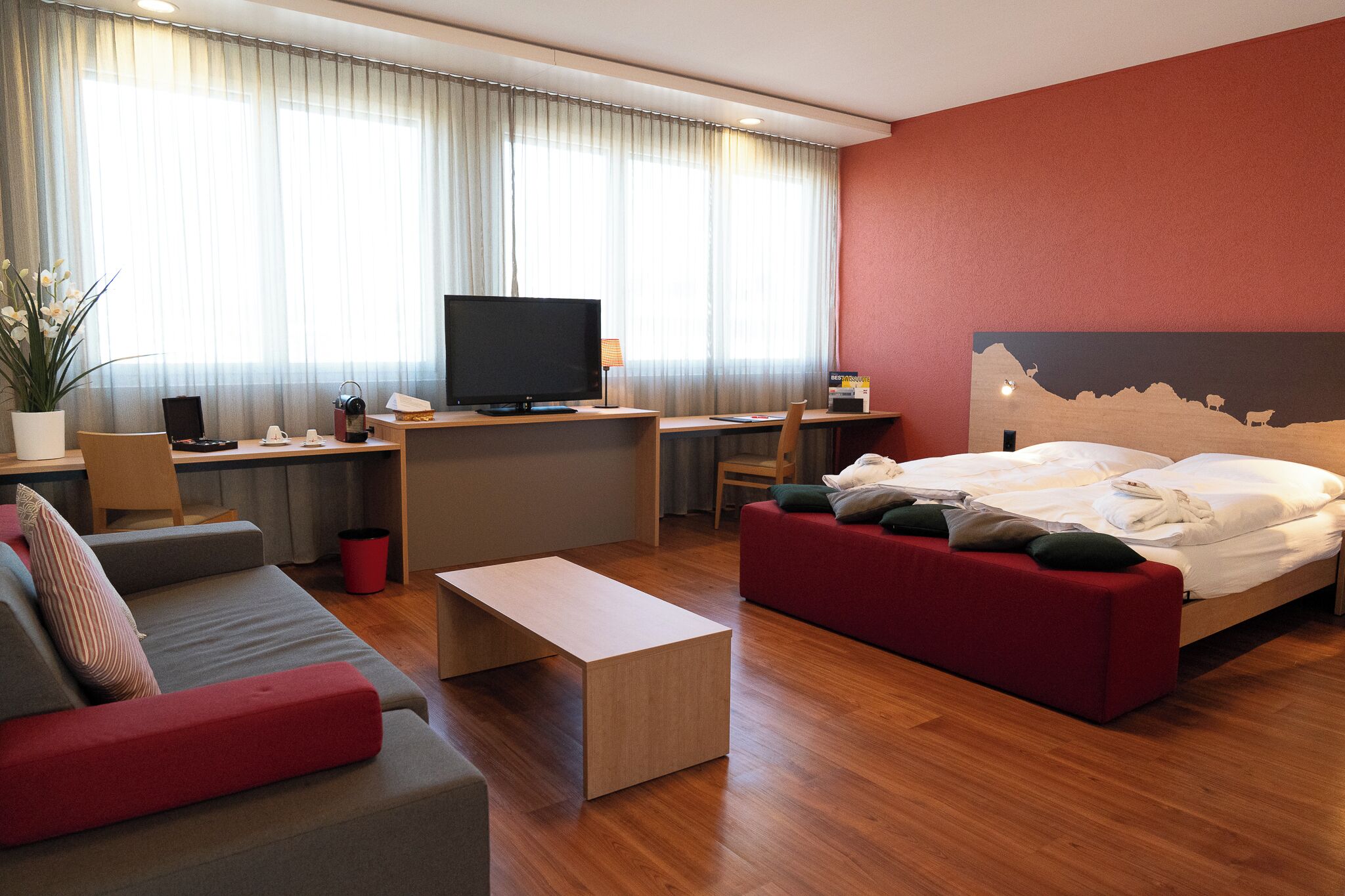 A modern hotel room with a bed, a desk with a computer, a television, a sofa, and a small table.