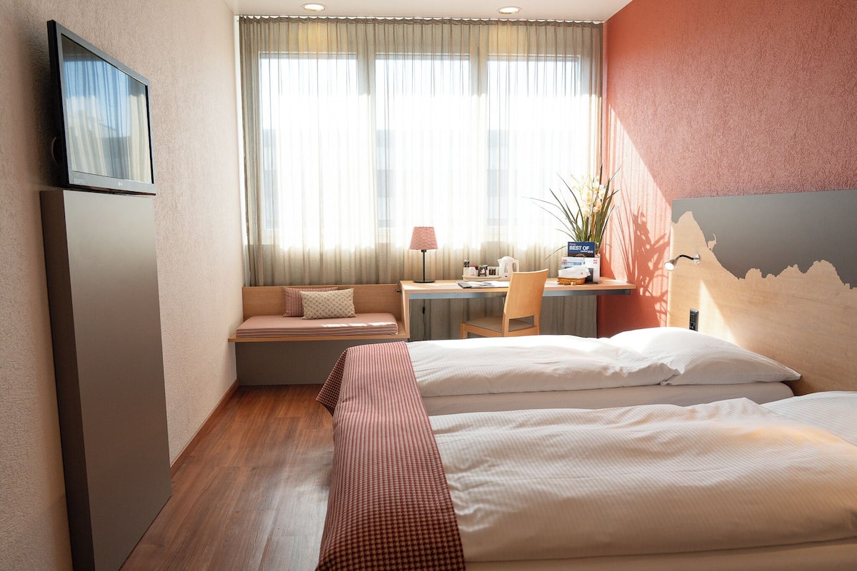 A hotel room with a bed, a desk, a chair, a TV, and a window with curtains.