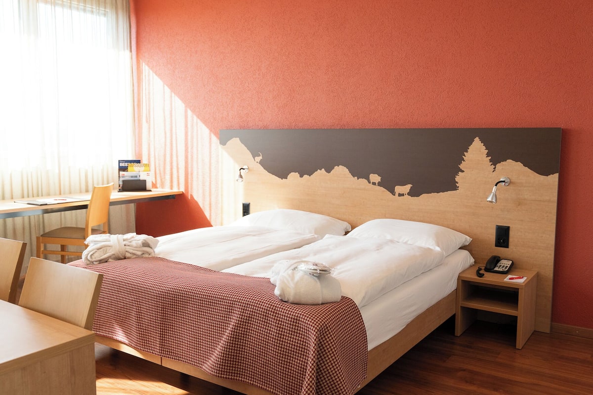 A hotel room with a bed, a desk, a chair, and a nightstand with a phone.