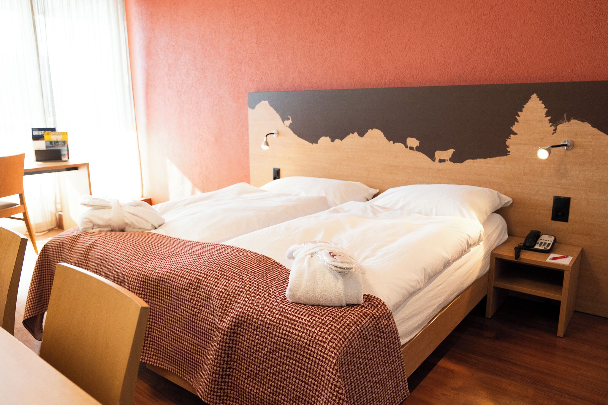 A hotel room with a bed, a bedside table with a phone, and a wall with a mountain landscape mural.