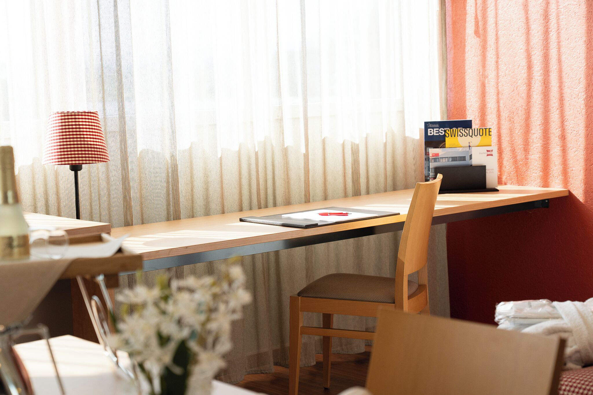 A hotel room with a wooden desk, a laptop, a red lamp, and a book titled 'BESSWISSQUOTE'.