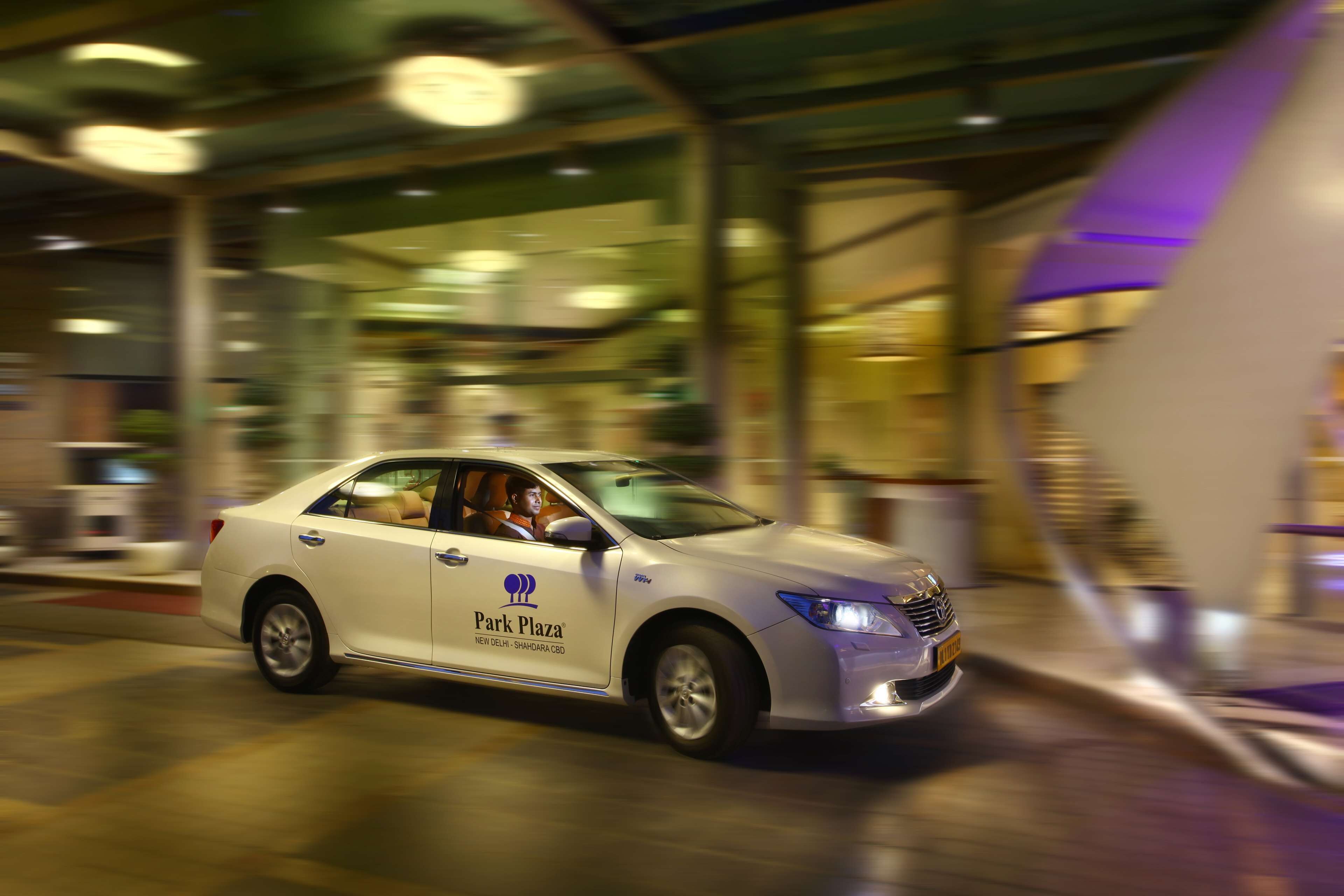 A white car with 'Park Plaza' branding parked in front of a building with large glass windows and a purple accent wall.