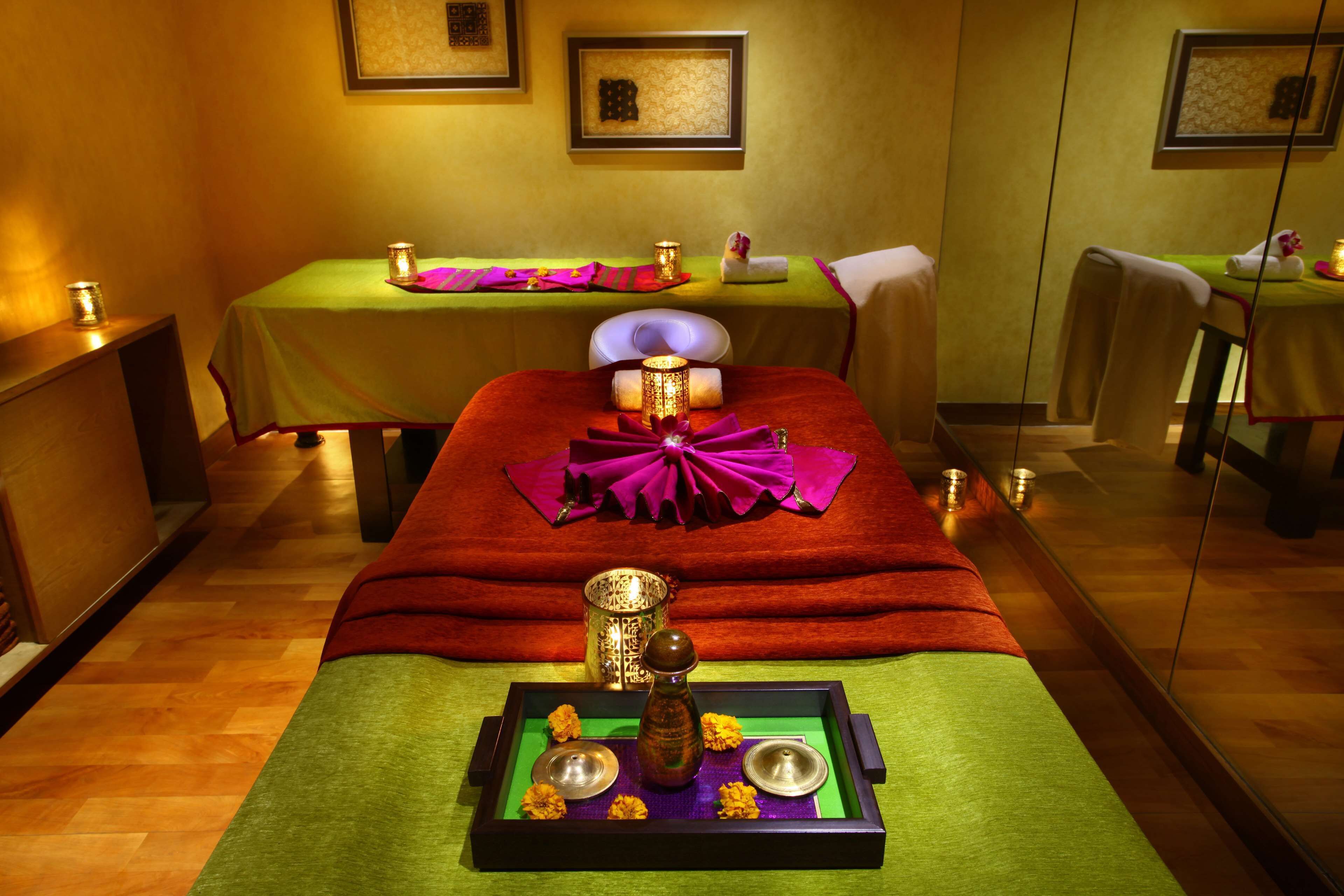 A spa room with two massage tables, each with a green tablecloth and a red blanket. There are candles and a decorative tray with flowers and a small bowl on the tables.