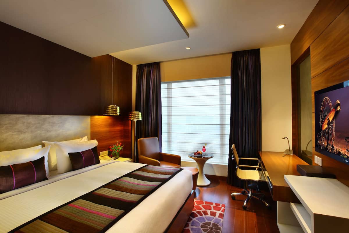 A hotel room with a large bed, a desk, a chair, a small table with a drink, and a TV on the wall.