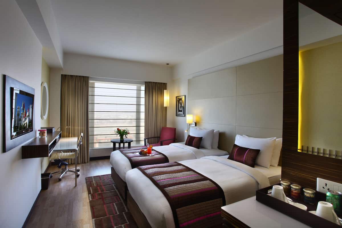 A hotel room with two beds, a desk, a chair, a small table with a drink, and a window with curtains.