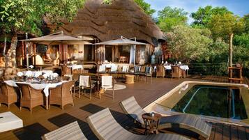 An outdoor dining area with wicker furniture and tables, a pool, and a thatched-roof structure.