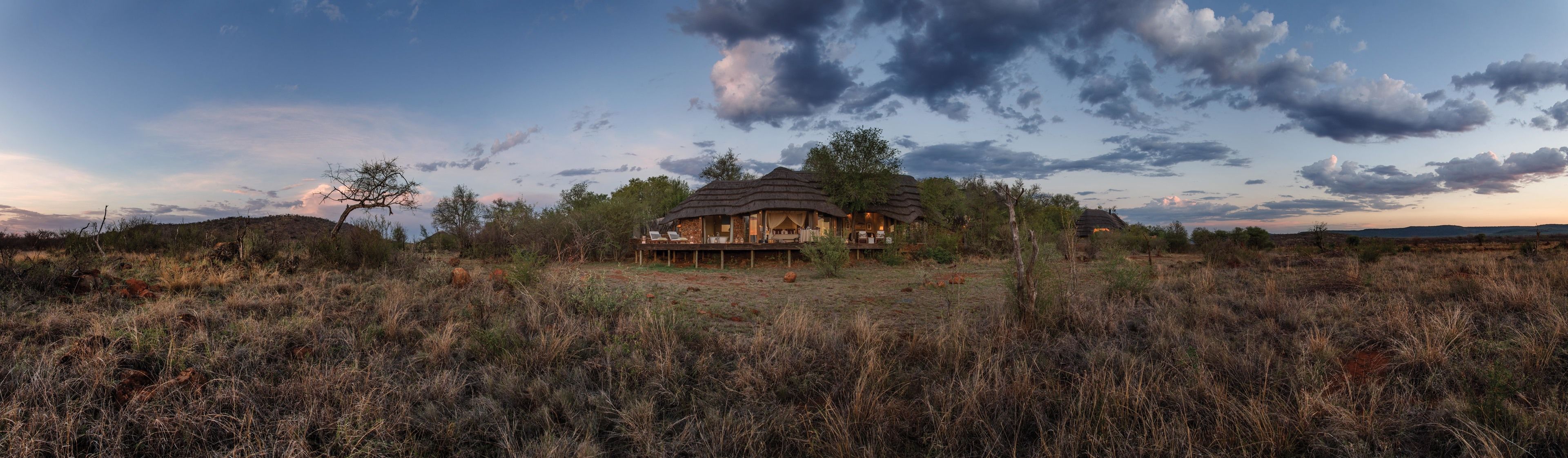 A thatched-roof structure with a deck and lounge chairs, surrounded by dry grassland and trees, under a partly cloudy sky.