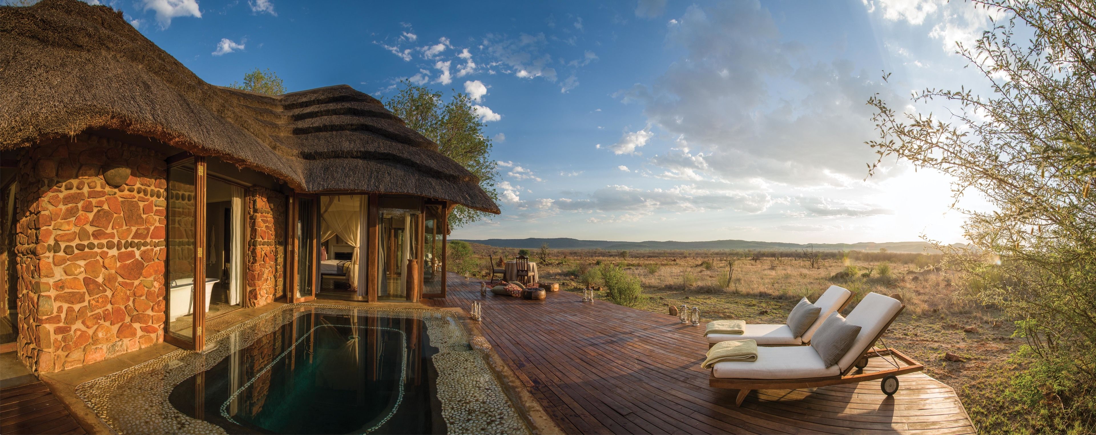 Un bâtiment à toit de chaume doté d’une piscine et de chaises longues, offrant une vue sur un paysage de savane.