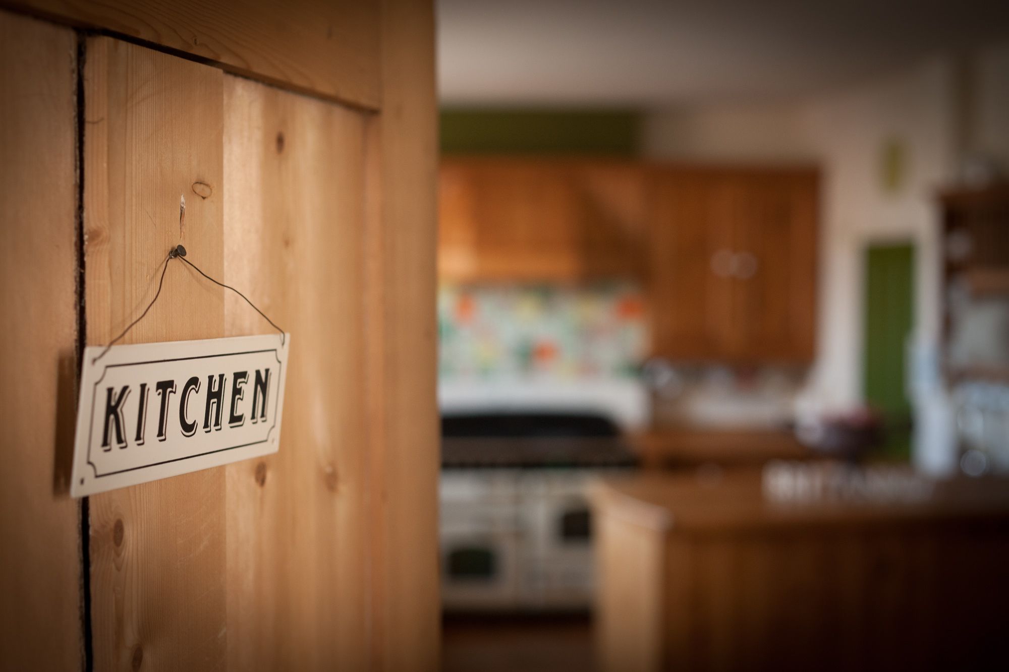 A wooden door with a sign reading "KITCHEN" and a blurred view of a kitchen area with wooden cabinets and appliances.