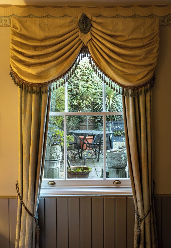 A window with a decorative valance and curtains, a view of an outdoor patio with a table and chairs, and a stone planter with plants. - Britannia House (Lymington)