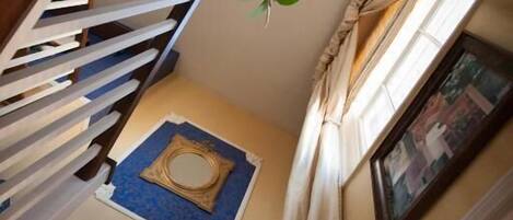 A staircase with a white railing, a blue mat, a framed mirror, and a window with curtains.