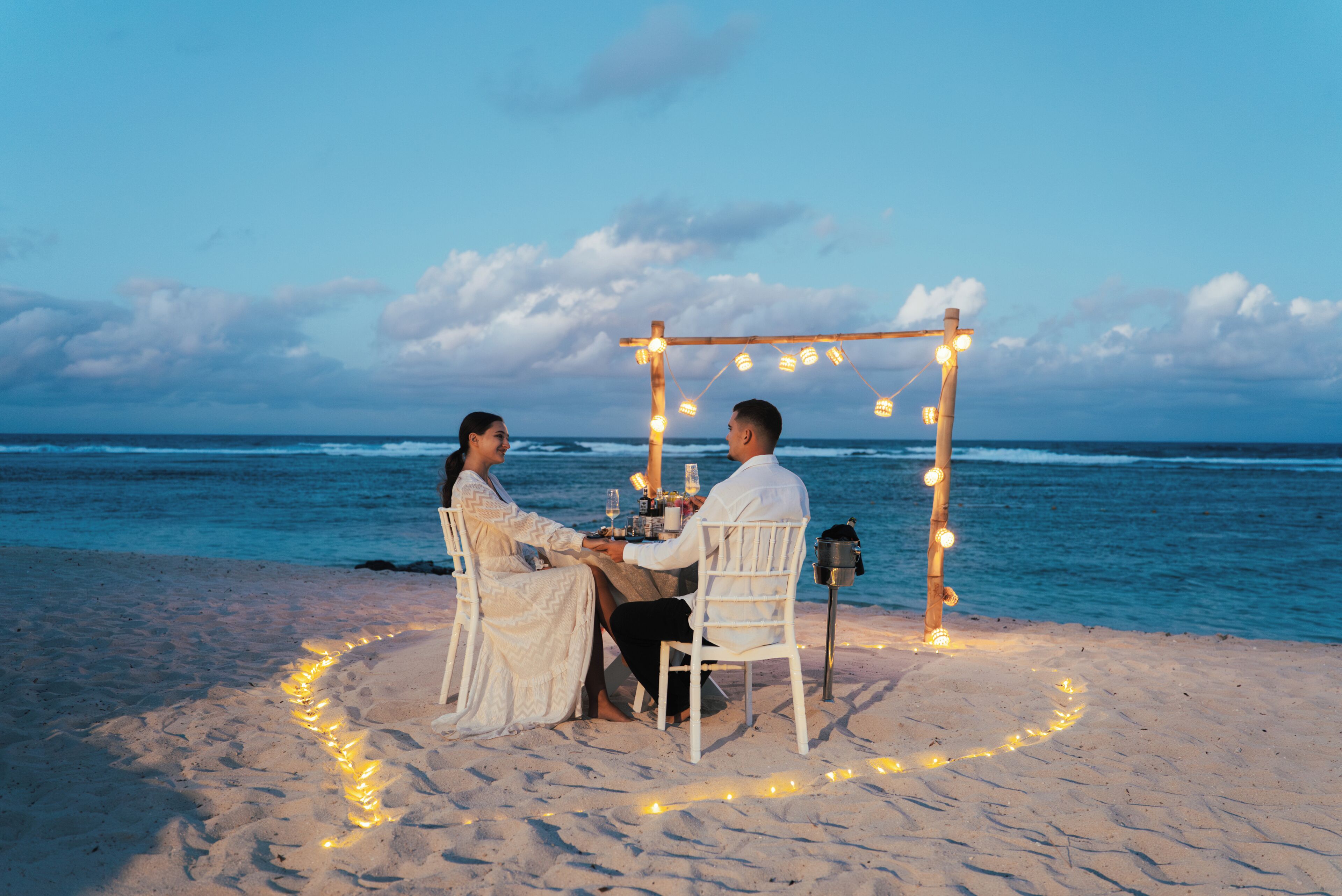 A couple dining at a beachside table with a wooden arch overhead, surrounded by string lights.