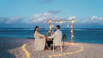 A couple dining at a beachside table with a wooden arch overhead, surrounded by string lights.
