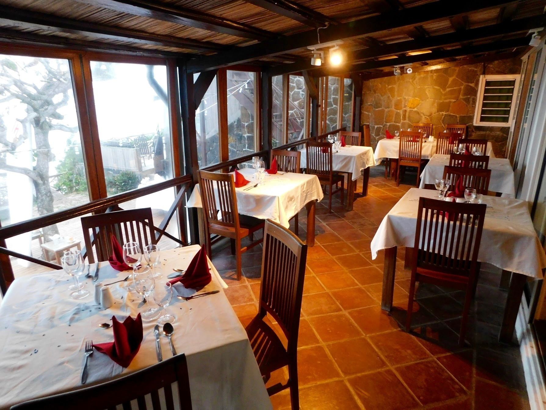Ein Restaurant mit mehreren Tischen, jeder Tisch ist mit einer weißen Tischdecke und roten Servietten gedeckt. Der Raum hat große Fenster und eine Steinsäule.