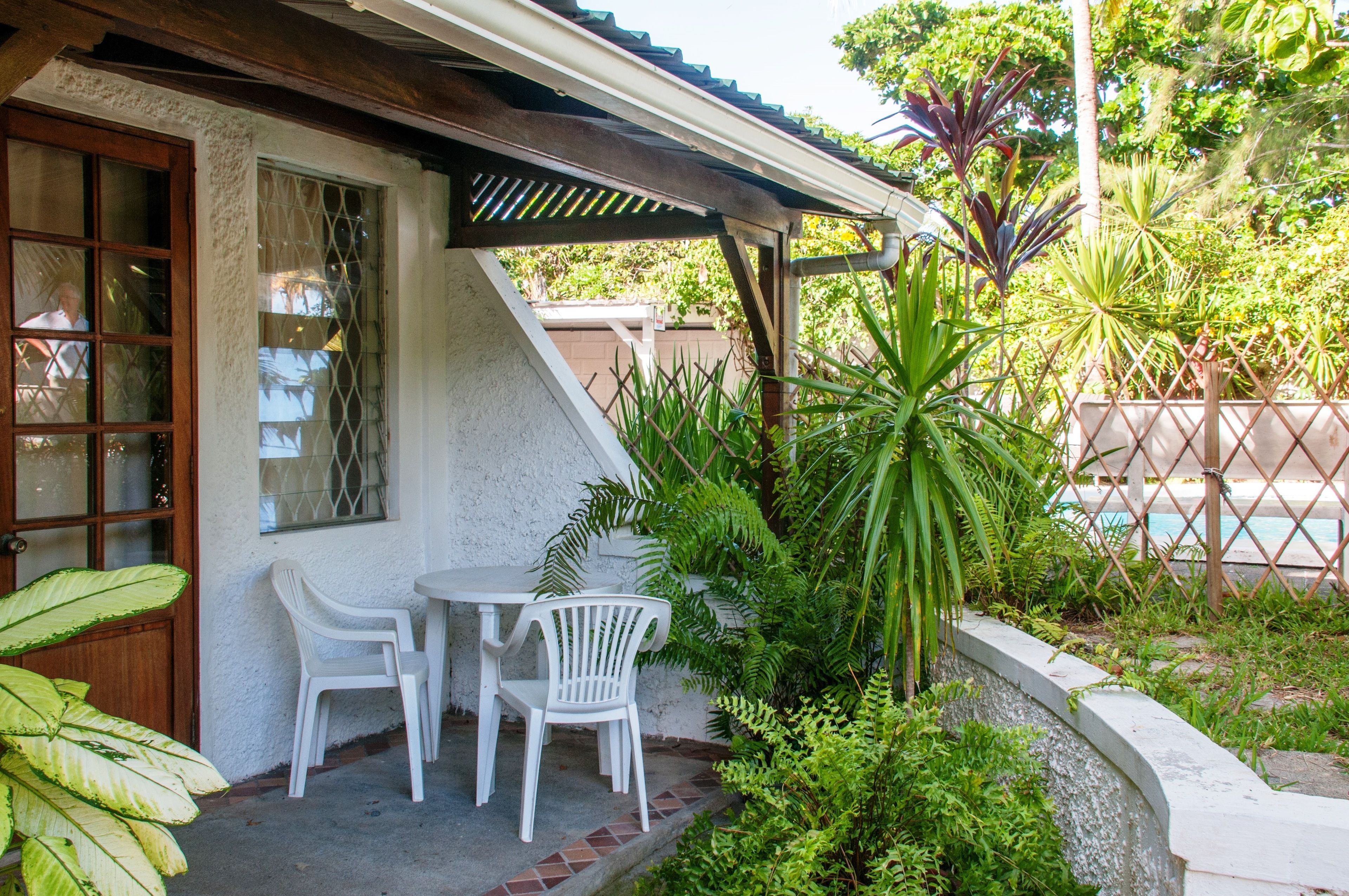 A patio with two white chairs and a table, surrounded by lush greenery and a wooden fence.