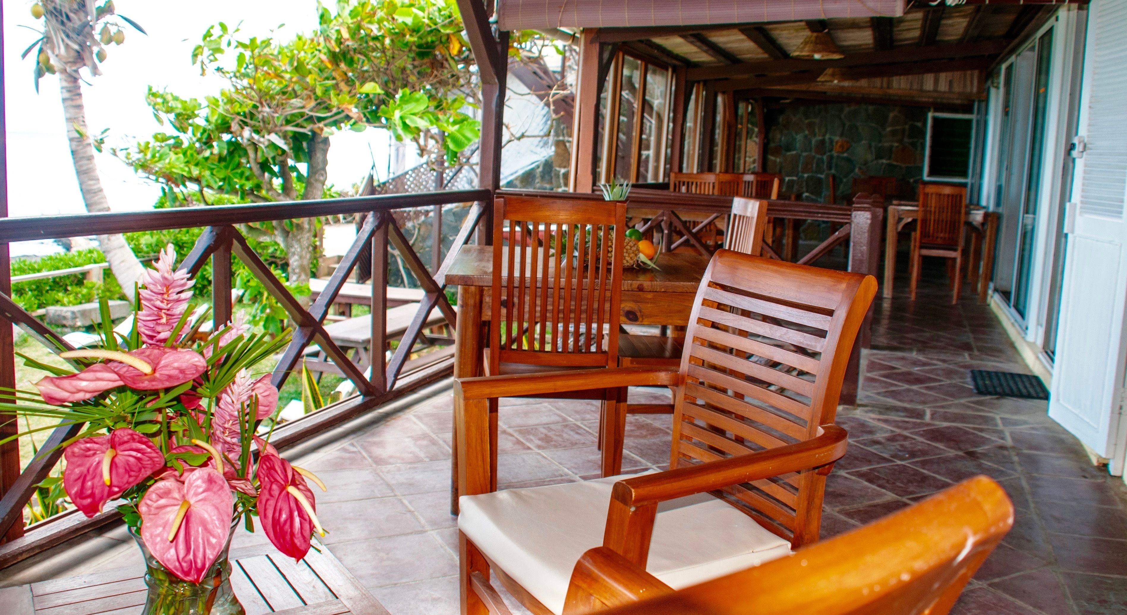 Una terraza de madera con una mesa y sillas, rodeada por una barandilla, y una planta en maceta con flores rosadas.