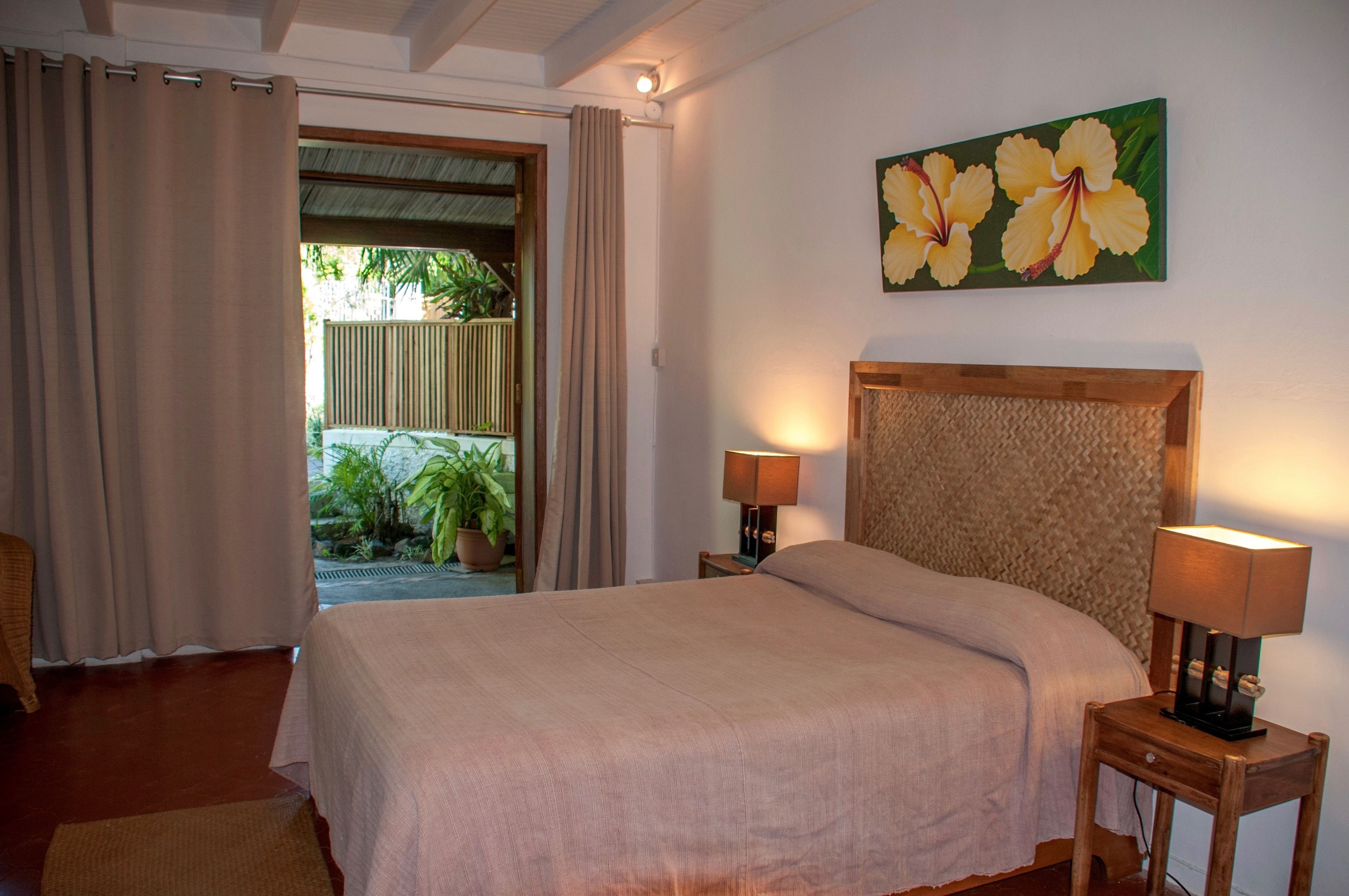 Une chambre à coucher avec un lit, deux lampes de chevet, une tête de lit en bois, un tableau représentant des fleurs et une vue sur un jardin par une porte ouverte.