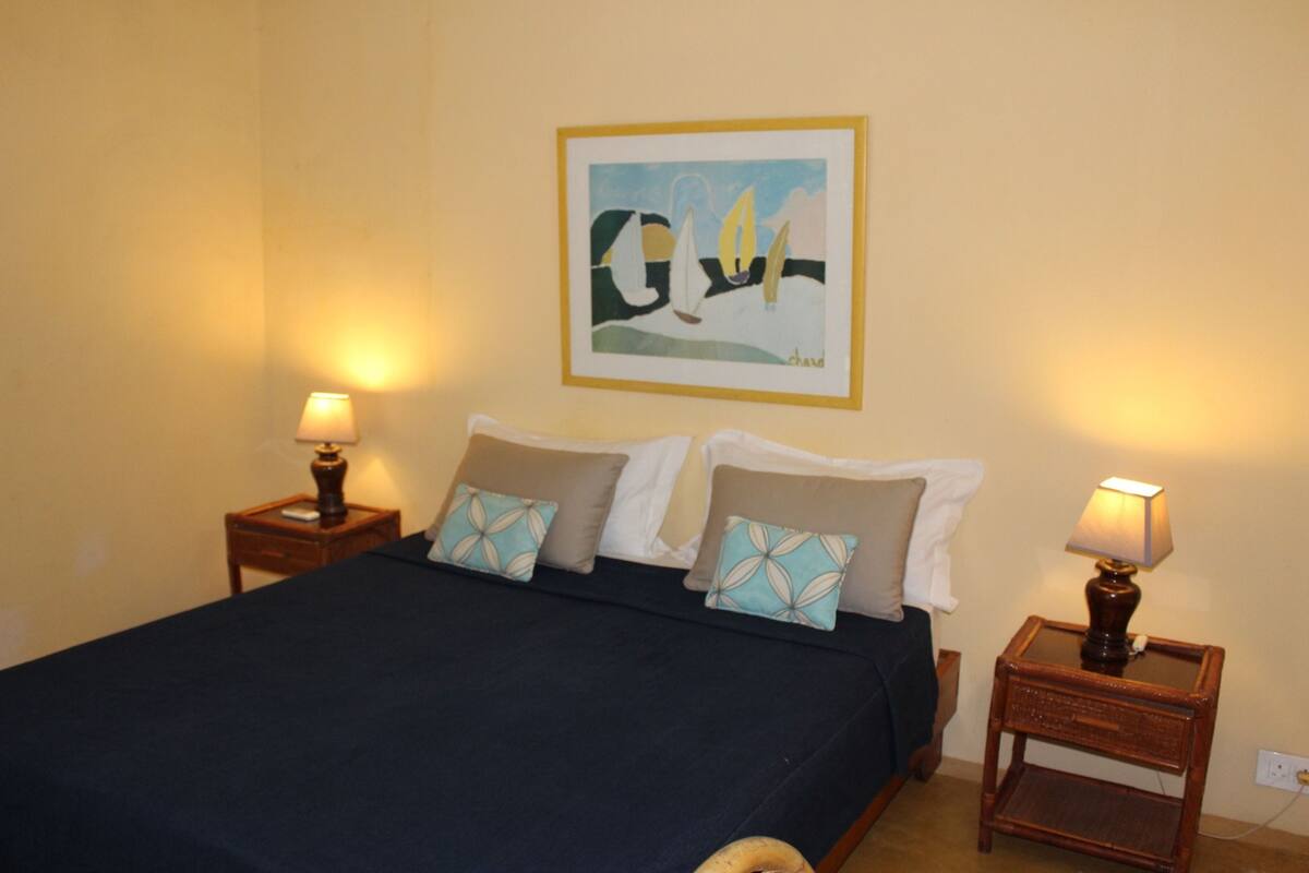 A neatly made bed with a dark bedspread and two decorative pillows, flanked by two bedside tables with lamps, a framed artwork on the wall, and a power outlet on the wall.
