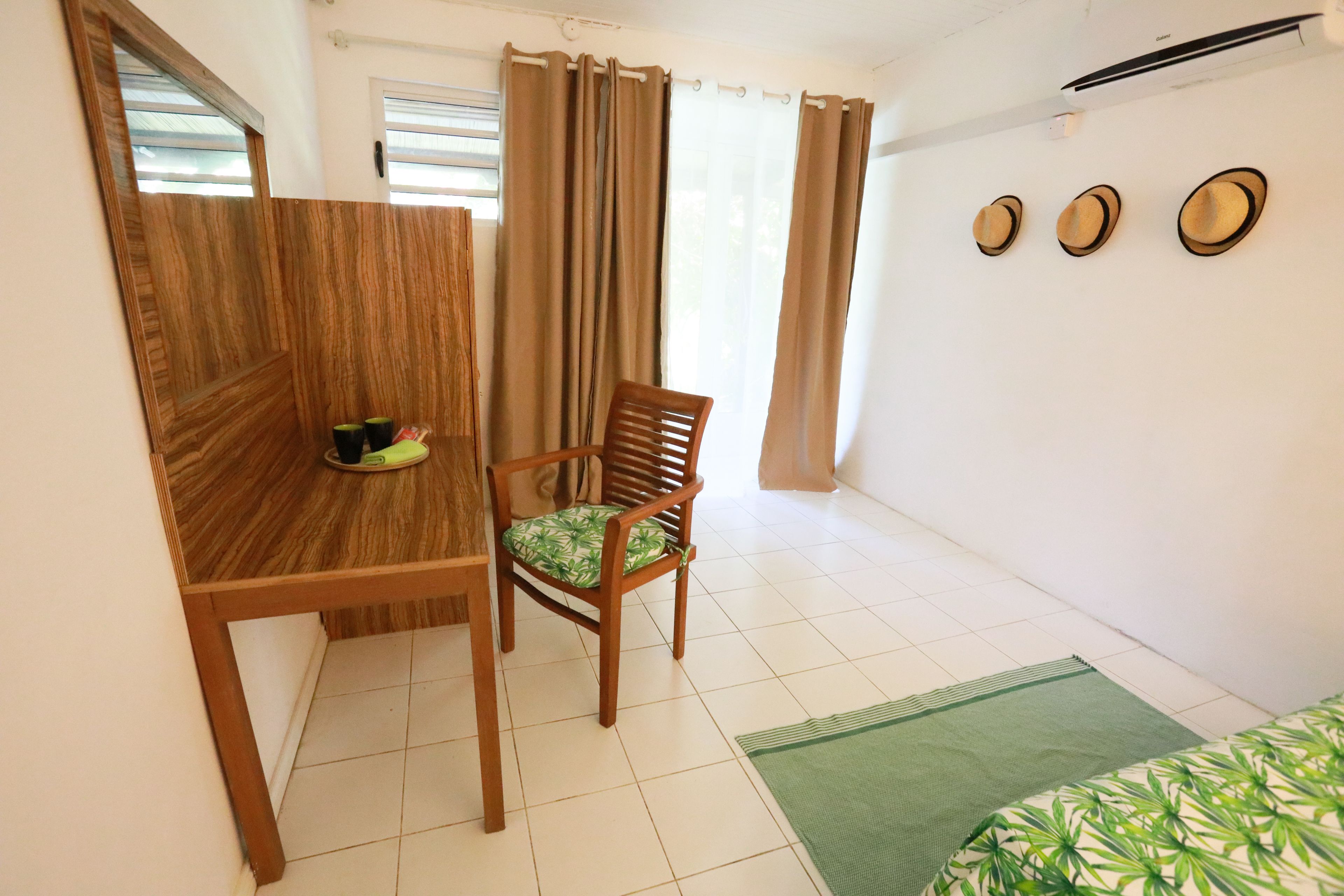 A room with a wooden desk, a chair with a green patterned cushion, a bed with a green and white patterned bedspread, and three wall-mounted hats.