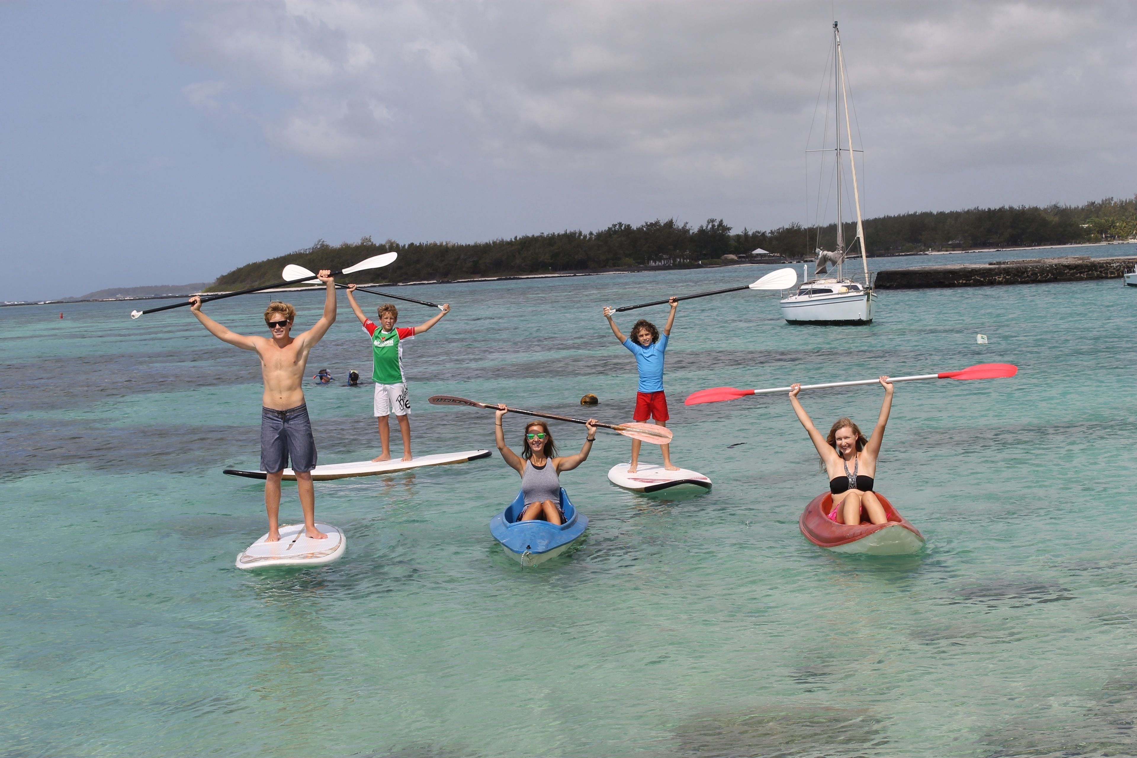 People standing on surfboards in a body of water with a sailboat in the background.
