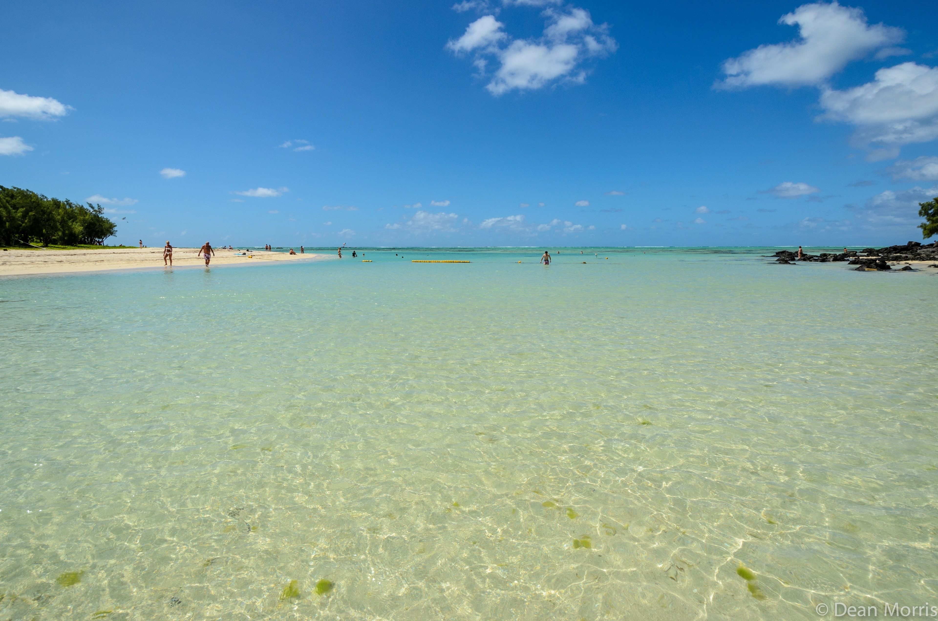 Una playa con aguas poco profundas y cristalinas, una orilla arenosa y algunas personas a lo lejos.