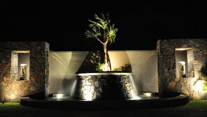 A nighttime outdoor water feature with a central tree, surrounded by stone walls and illuminated by ambient lighting.