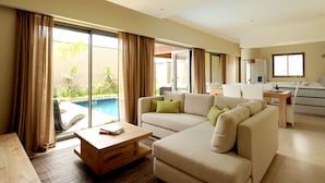 A modern hotel room with a large sofa, a wooden coffee table, and a view of a swimming pool.