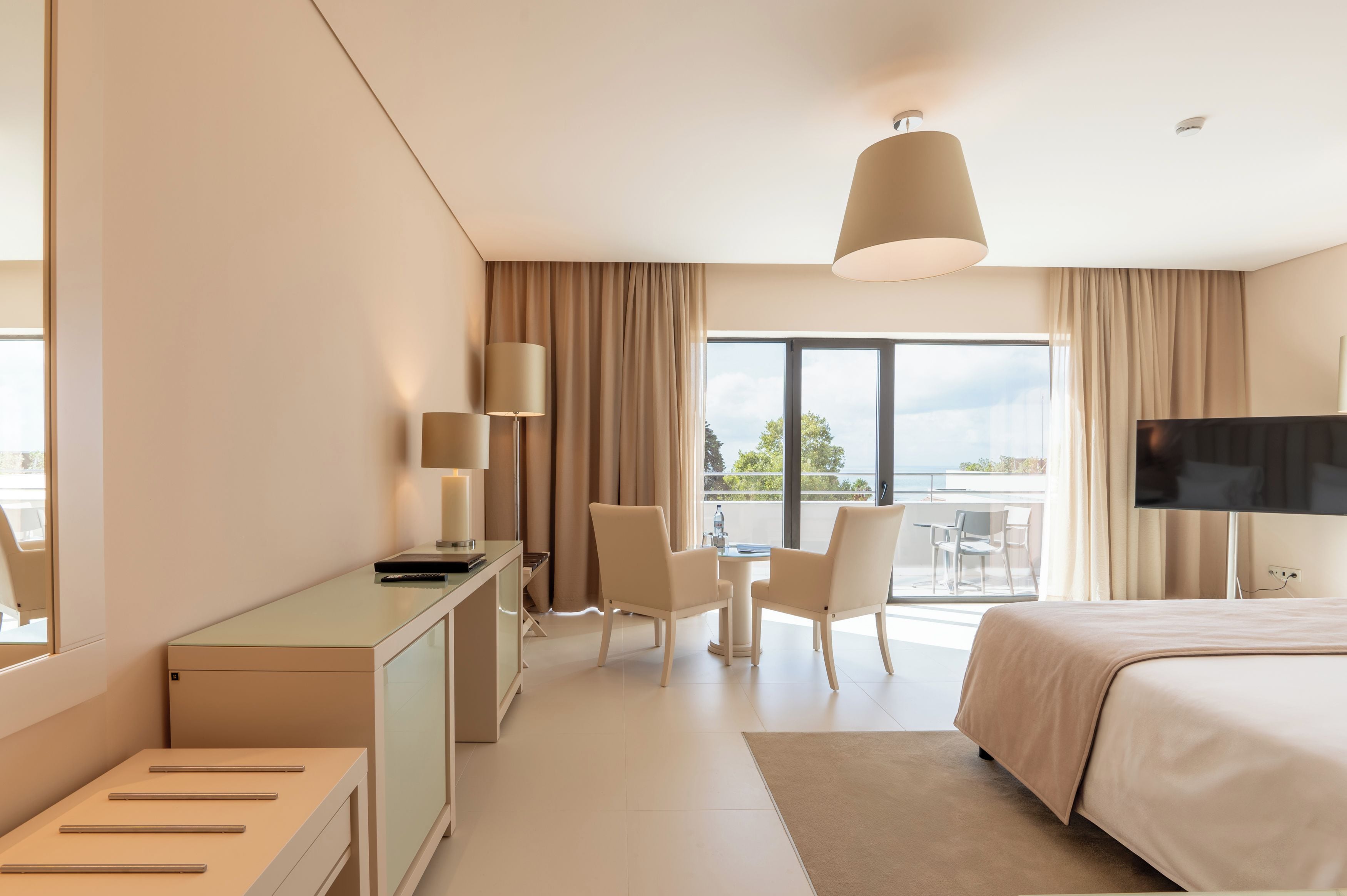 Habitación de hotel con cama, escritorio, sillas, televisor y balcón con vistas.