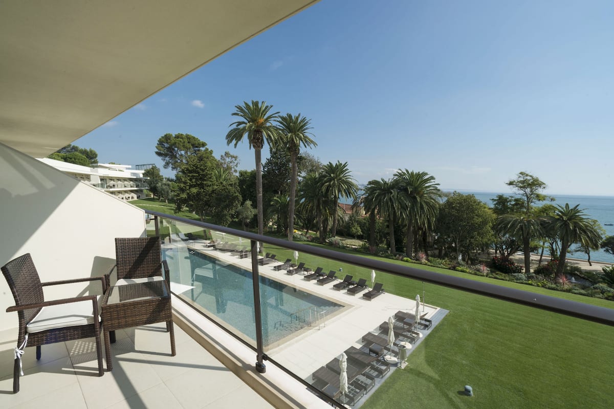 A balcony with a pool, lounge chairs, and a view of the sea and palm trees.