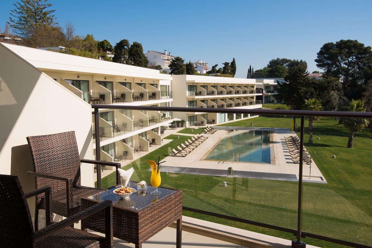 A balcony with wicker furniture, a glass table, and a view of a pool and hotel building.