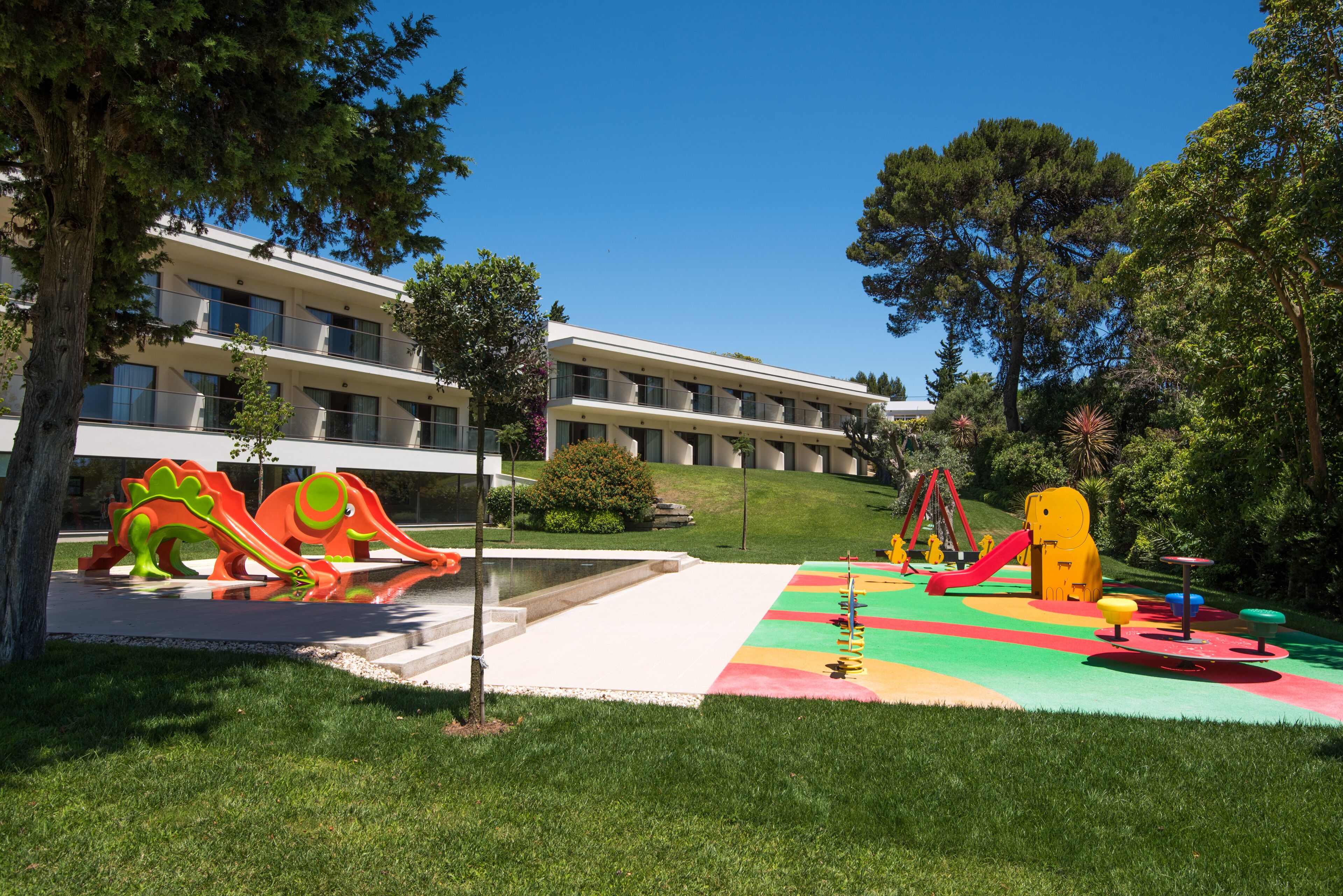A playground with colorful slides and climbing structures, surrounded by a building with multiple balconies and a green lawn.