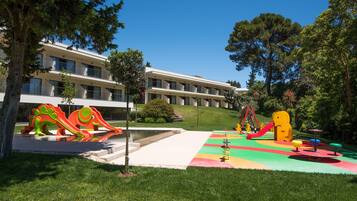 A playground with colorful slides and climbing structures, surrounded by a building with multiple balconies and a green lawn.