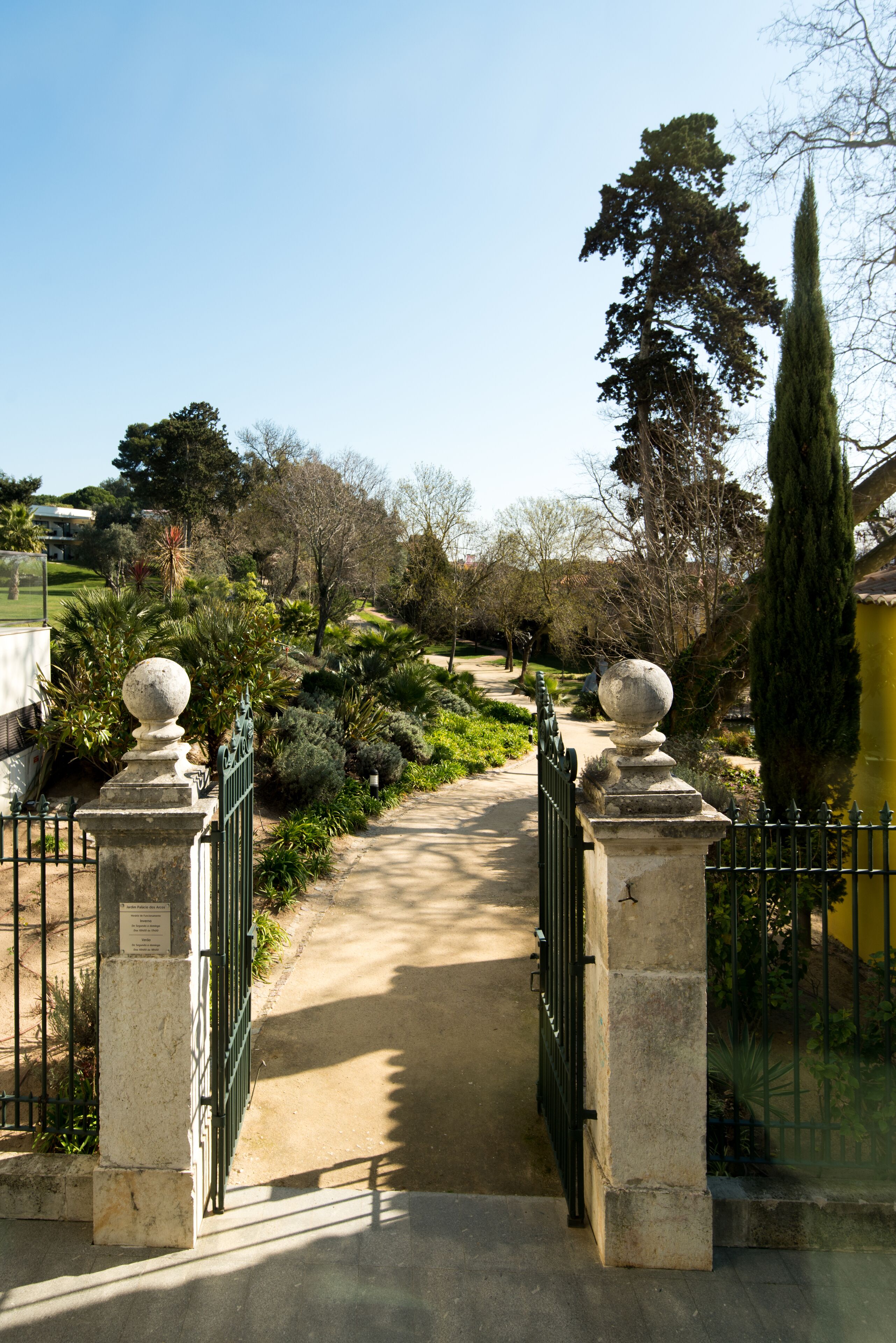 Uma passagem serpenteia por um jardim com um portão de pedra e uma árvore alta à direita.