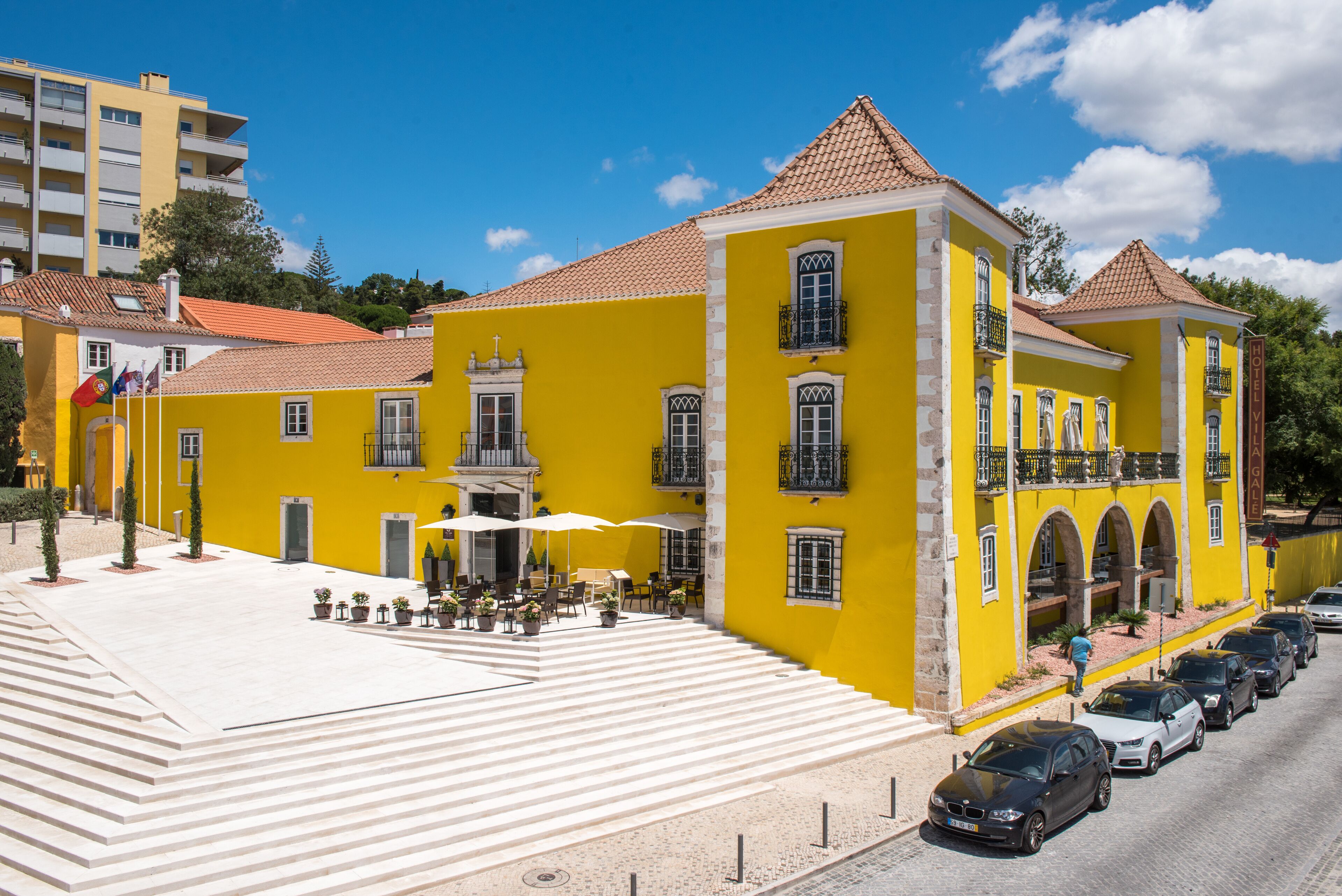 Un edificio amarillo con techo de tejas, zona de estar al aire libre y una escalera que conduce a la entrada.