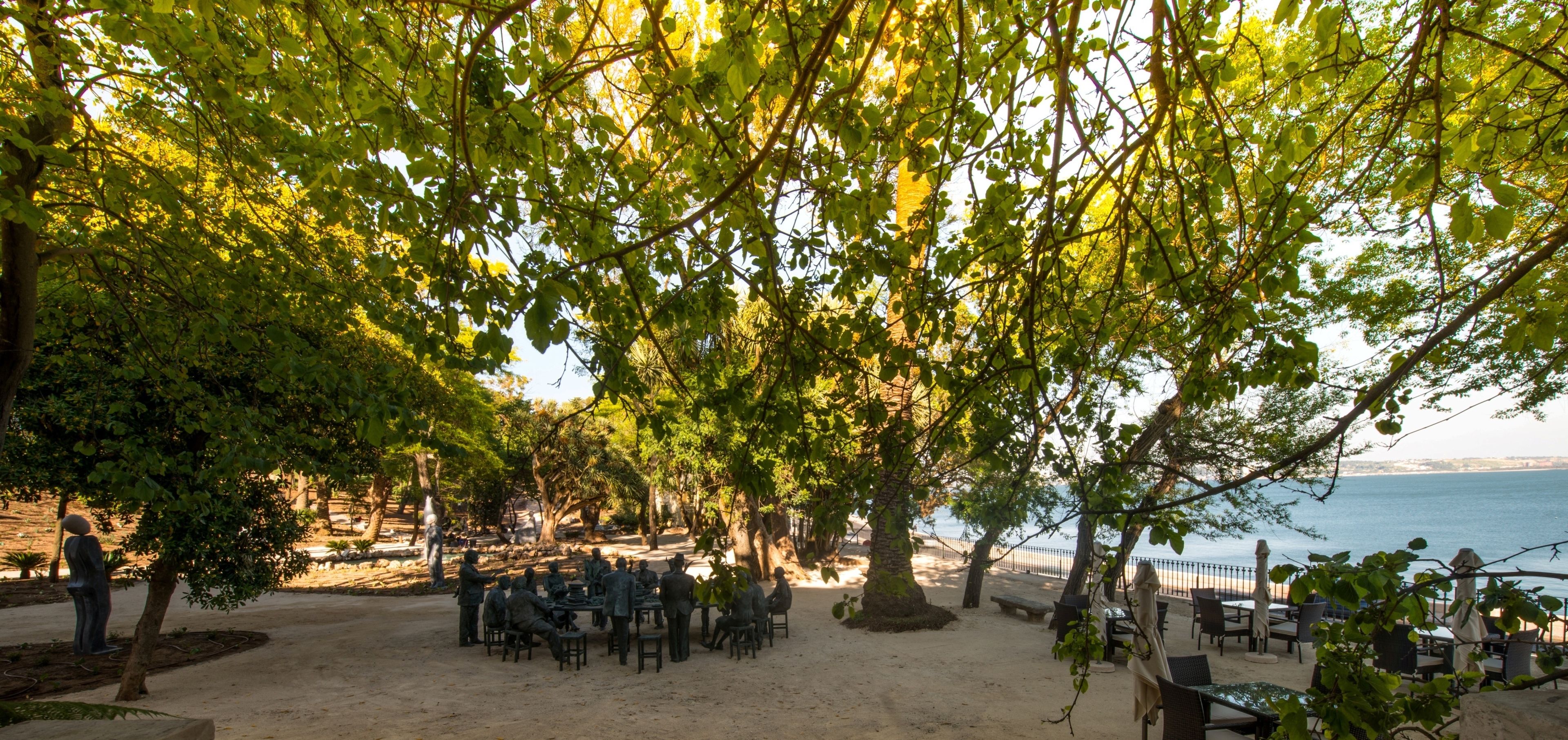 A beachside outdoor seating area with tables and chairs under trees, overlooking the sea.