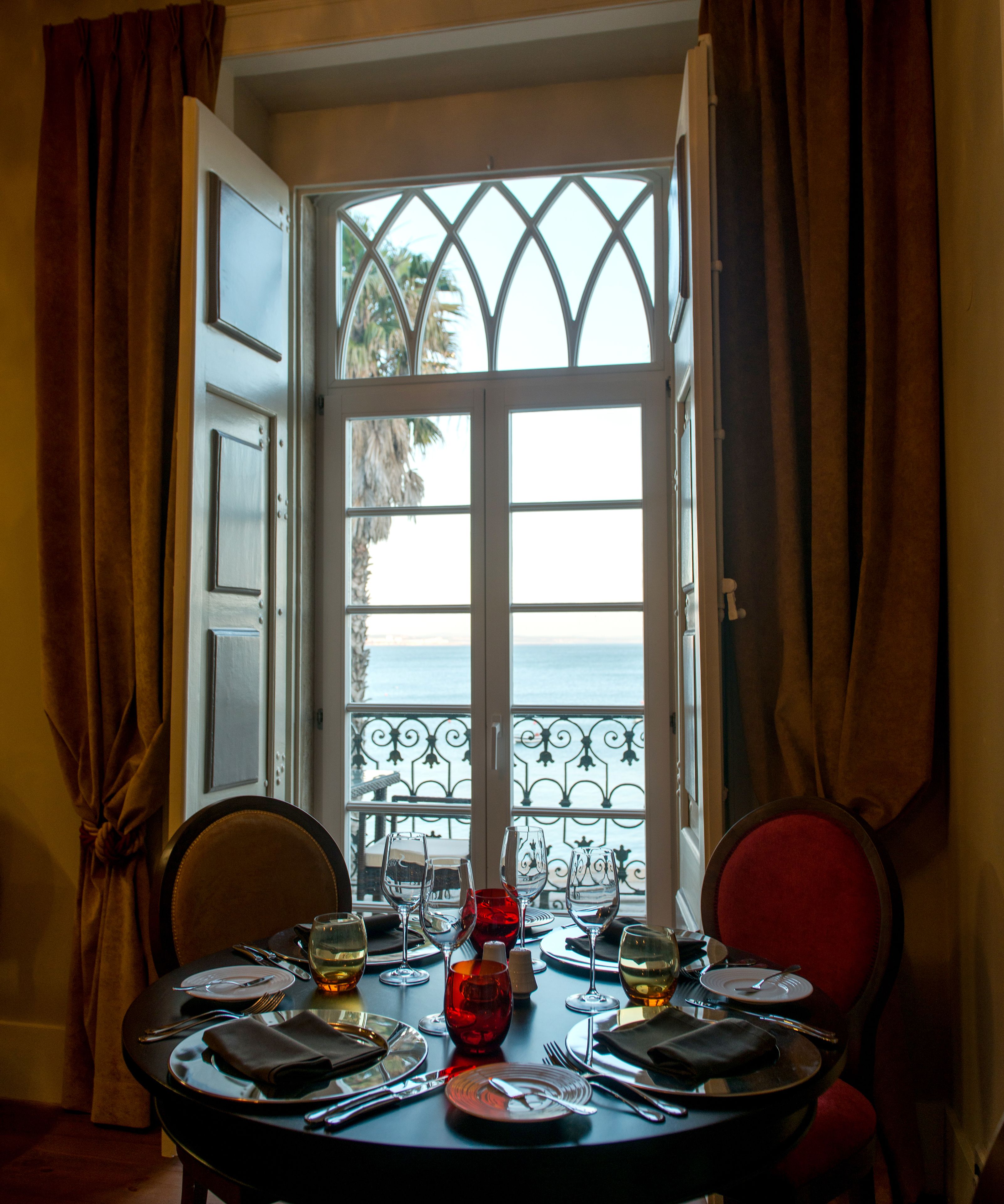 A dining table set for two with a view of the sea through a window with iron railings.