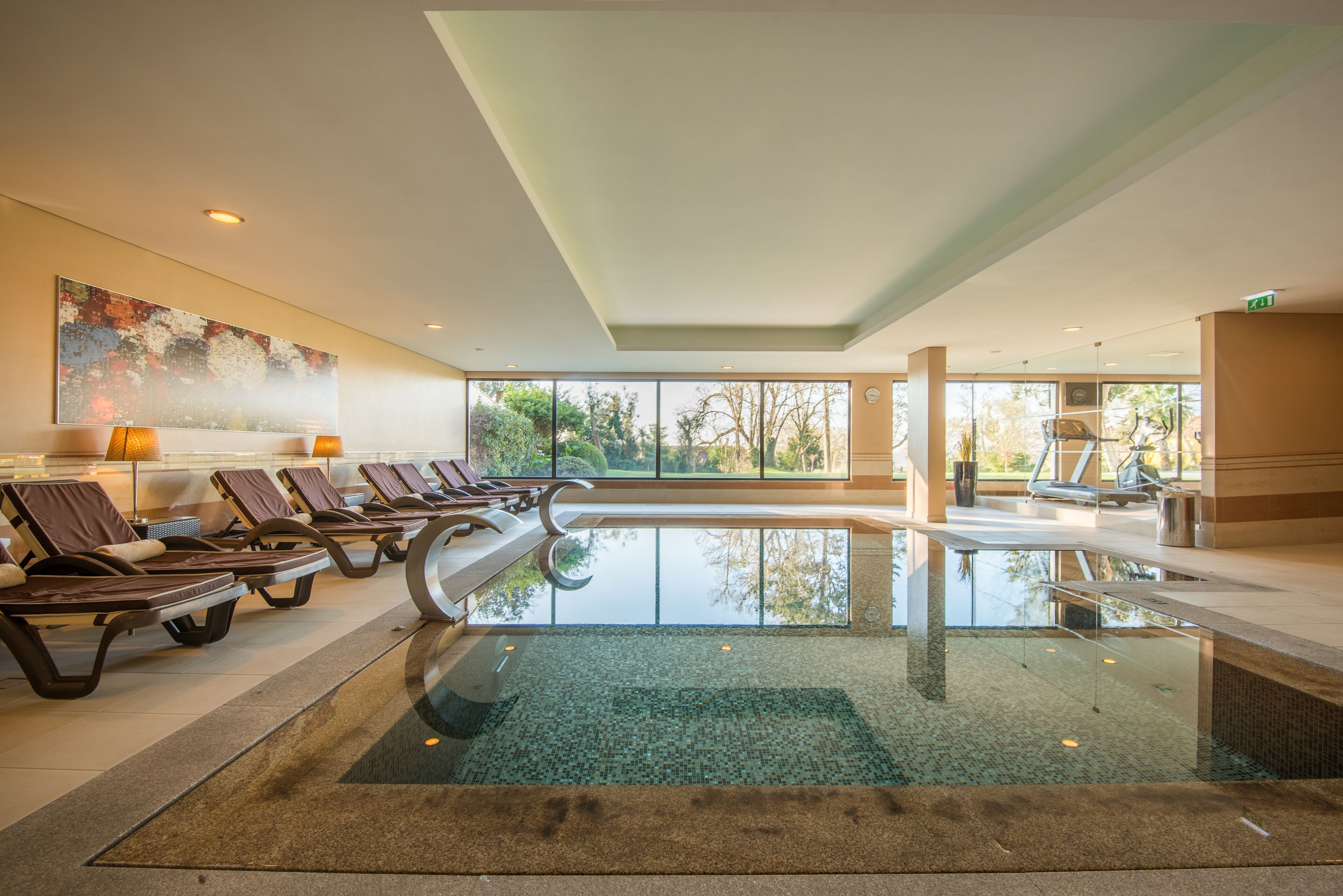 Un área de piscina interior moderna con sillones reclinables, una piscina de cristal y amplias ventanas que ofrecen vistas al exterior.