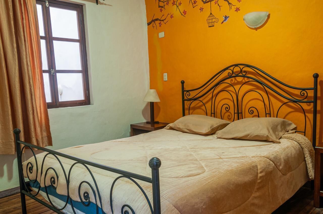 Ein Einzelbett mit Metallrahmen und beige bezogenen Bettwaren in einem Zimmer mit einer orangefarbenen Wand, einem Fenster mit Vorhängen und einer an der Wand befestigten Lampe.