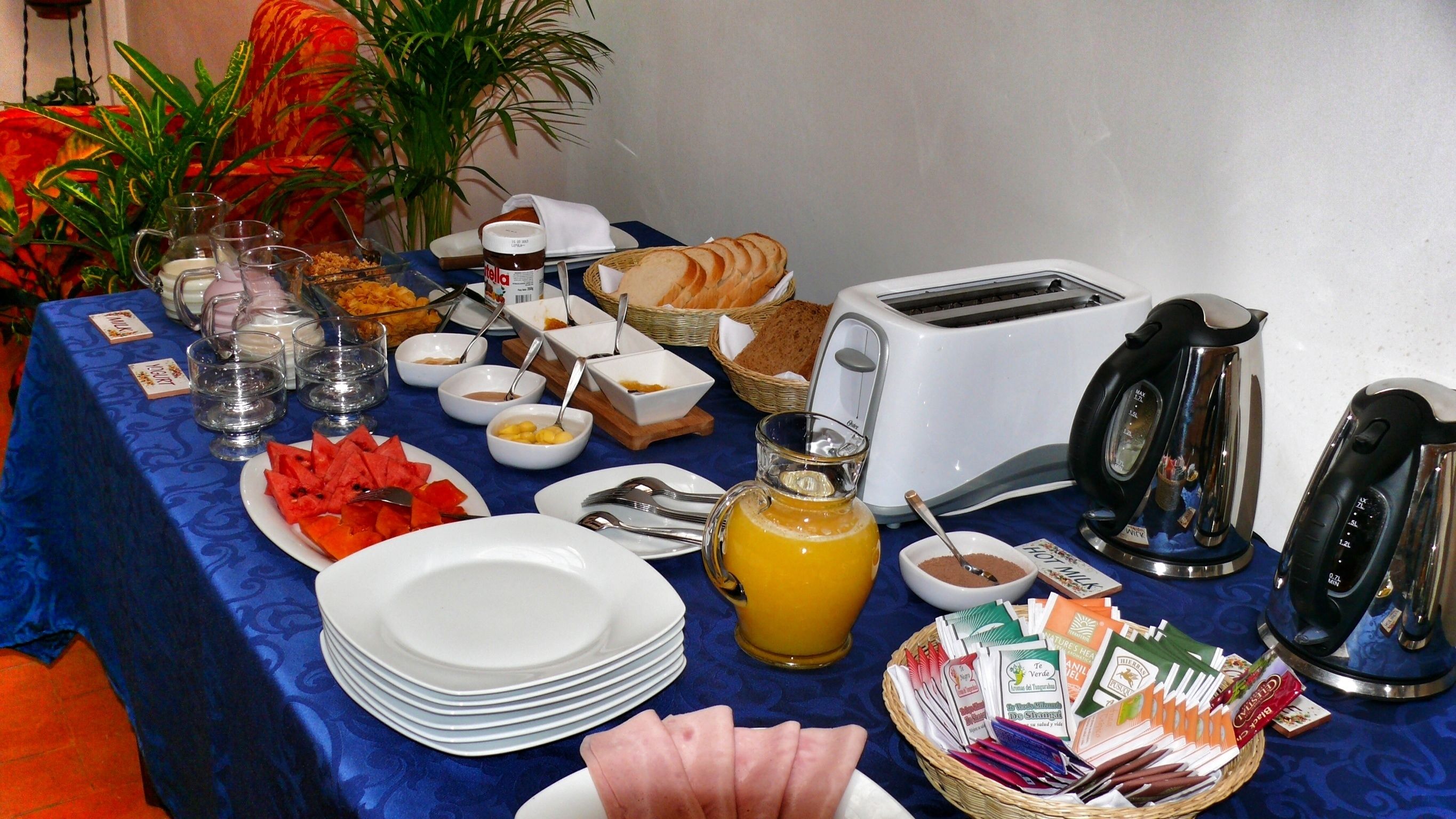A breakfast buffet with various food items, including bread, fruits, and condiments, arranged on a table with a blue tablecloth.