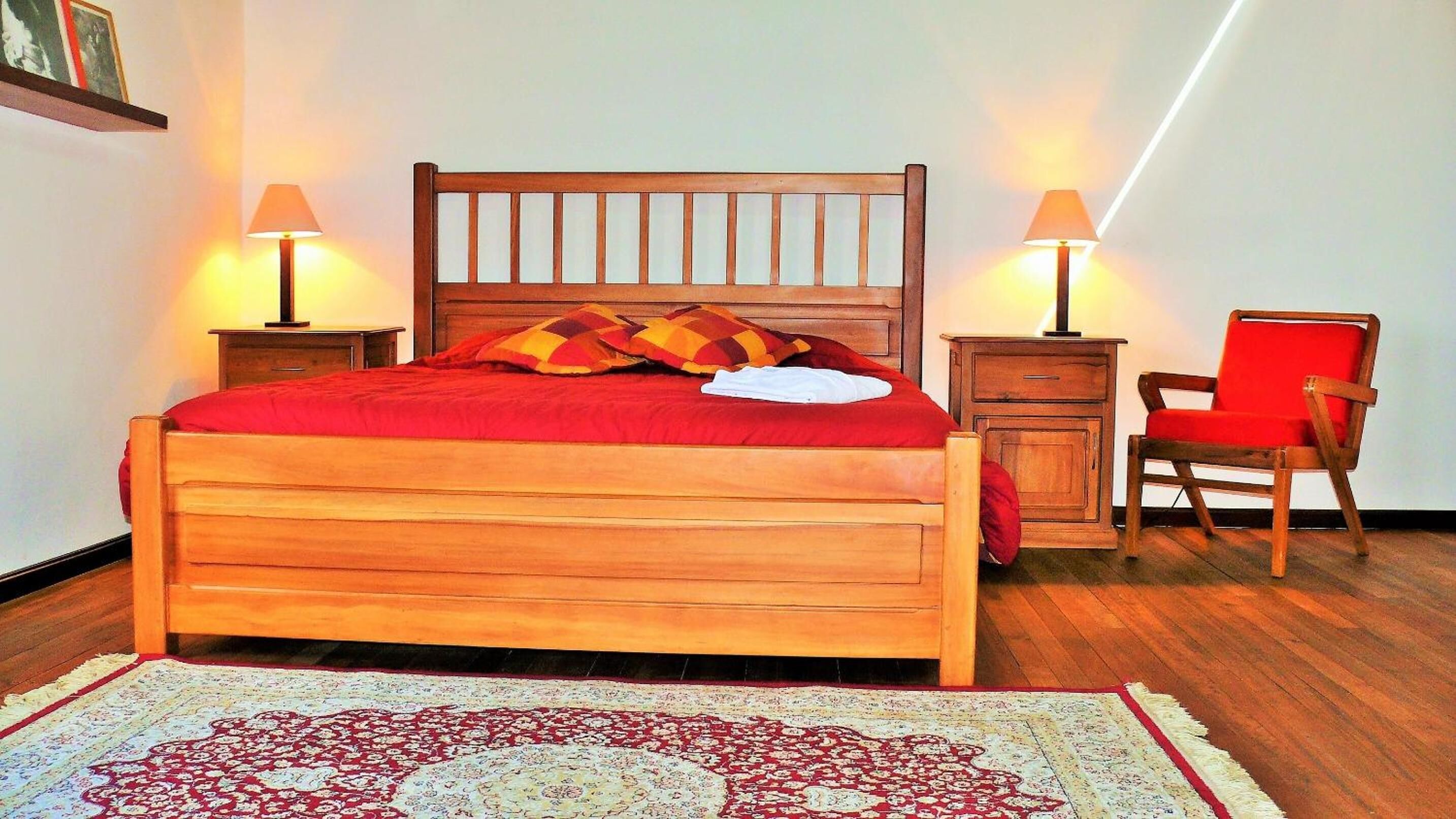 Una cama de madera con colcha y almohadas rojas, dos mesitas de noche con lámparas, una silla roja y una alfombra estampada.
