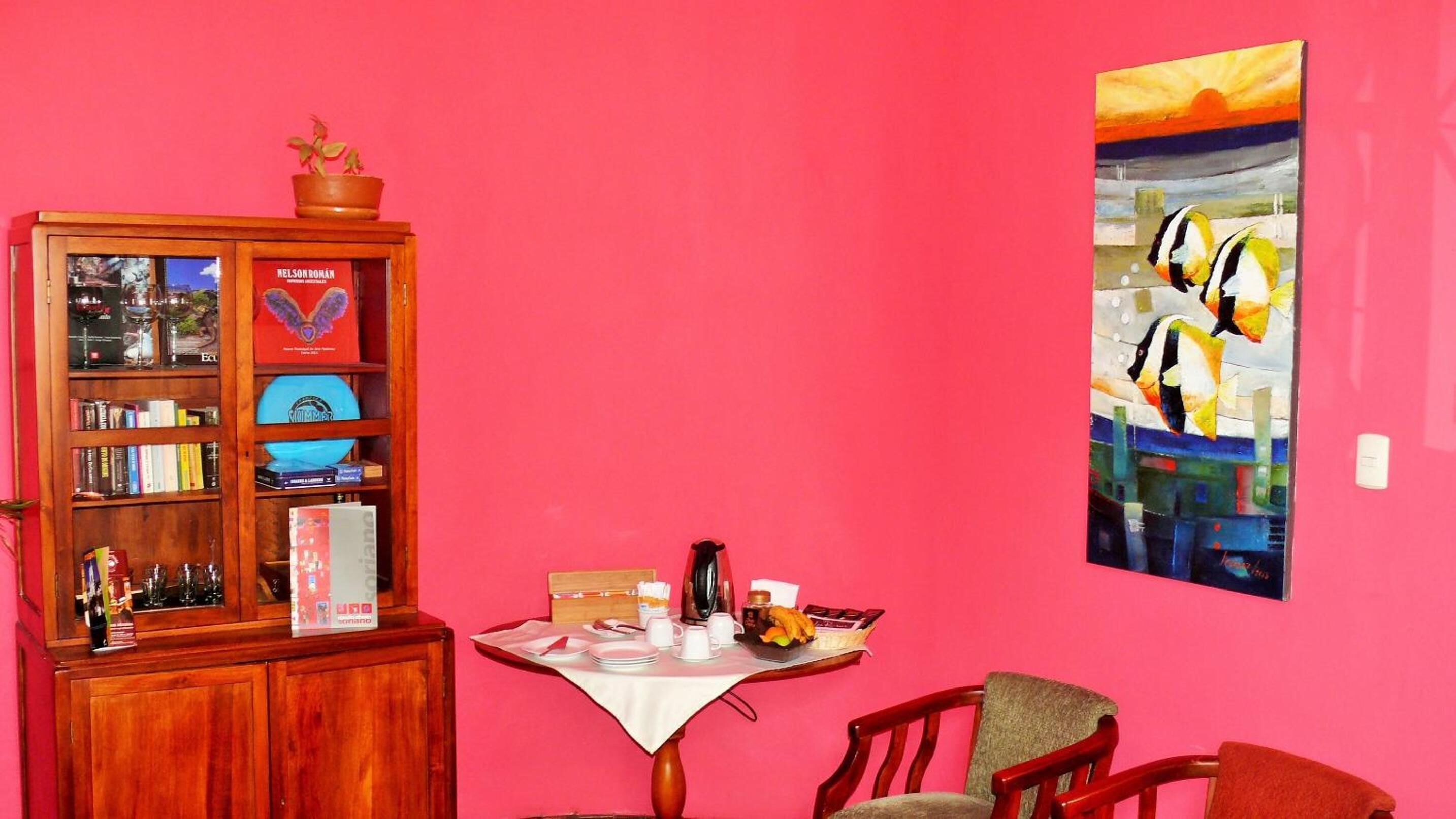 A room with a wooden cabinet filled with books and decorative items, a table with a white tablecloth and various objects, and a colorful painting on the wall.