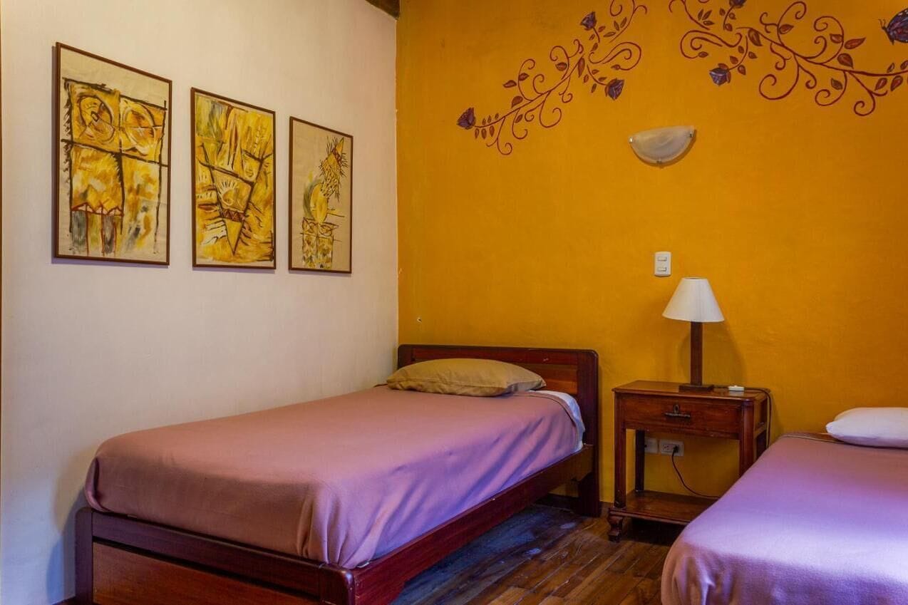 A room with two beds, a nightstand with a lamp, and three framed artworks on the wall.