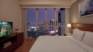 A hotel room with a large window offering a city view at night, a bed, a TV on a wooden console, and a nightstand with a lamp.