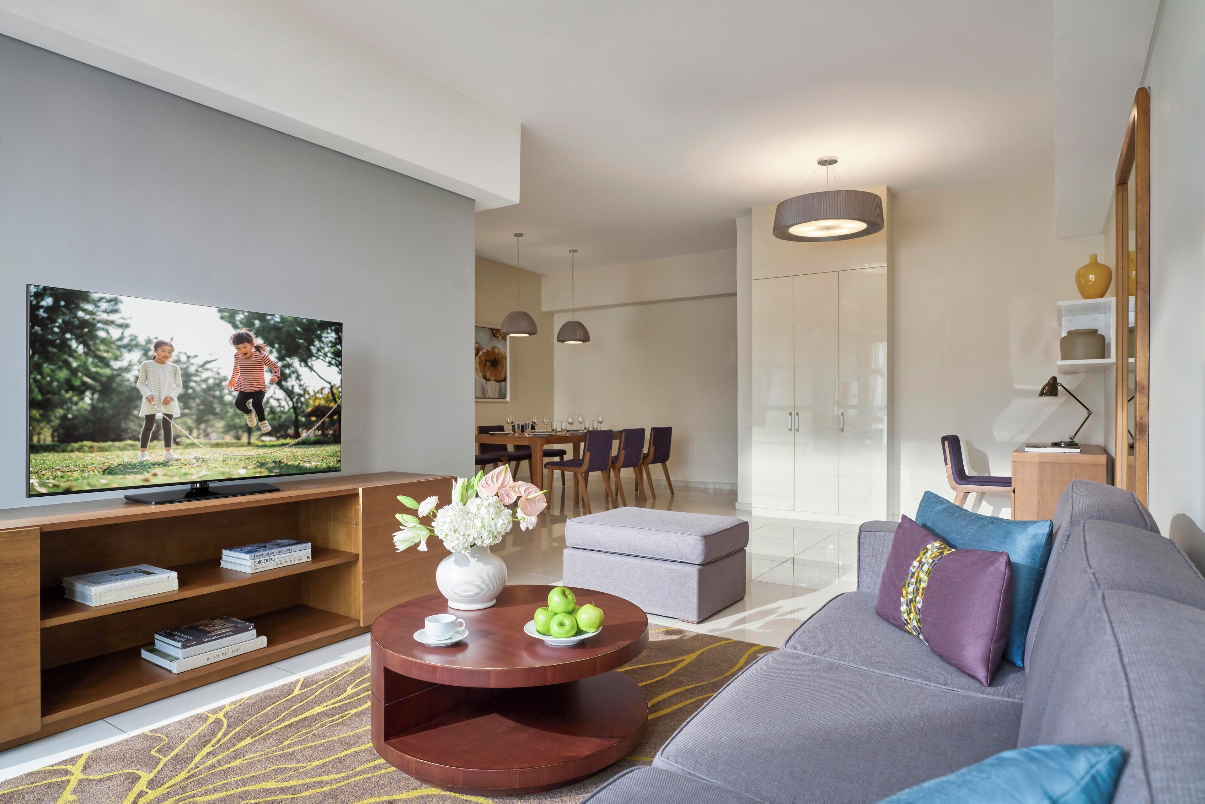 A modern living room with a television, a coffee table with books and flowers, a grey sofa with cushions, and a dining area in the background.