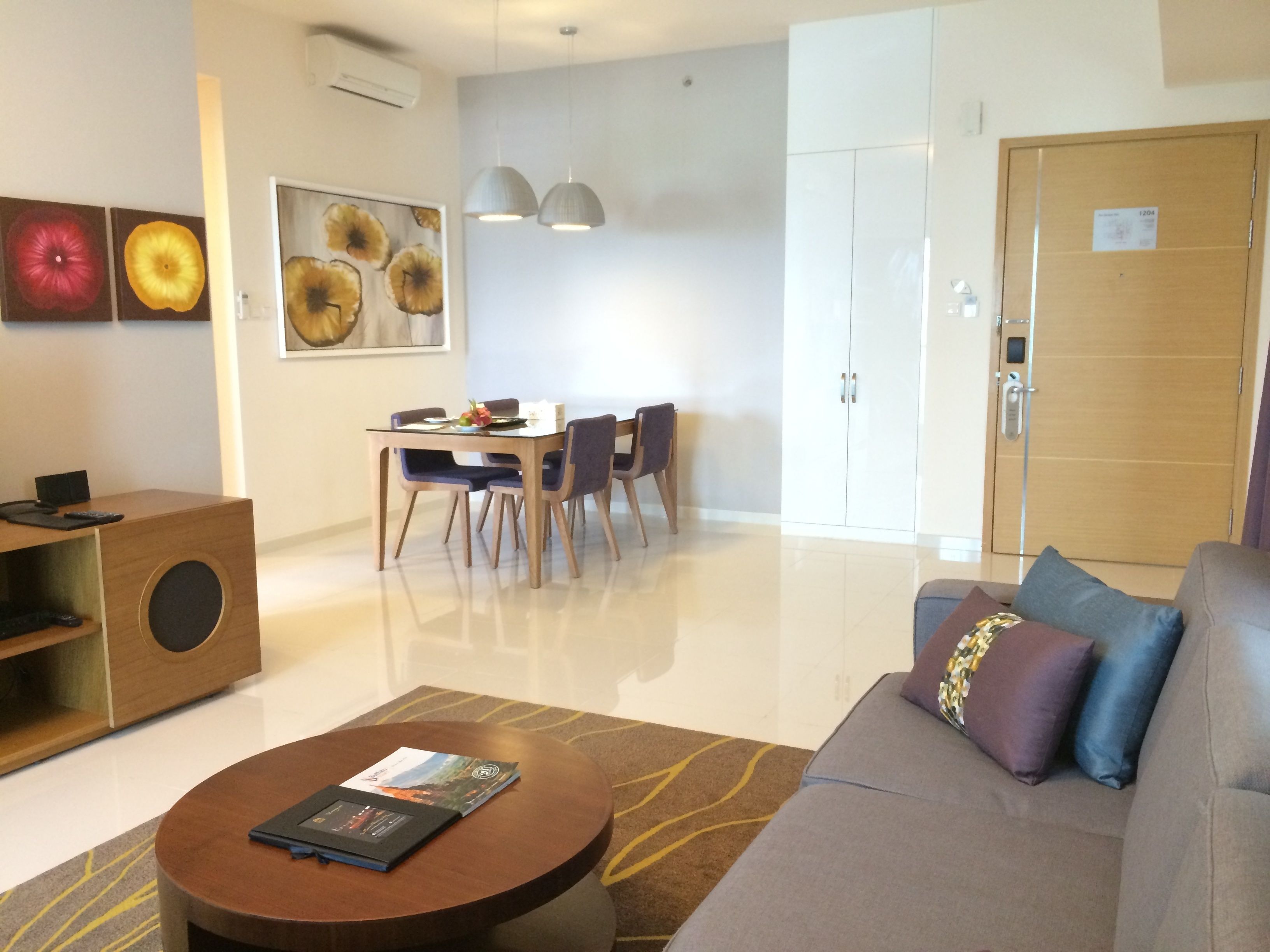 A modern living room with a sofa, coffee table, dining table, and chairs. There is a cabinet with a speaker, artwork on the walls, and a door with a number sign.